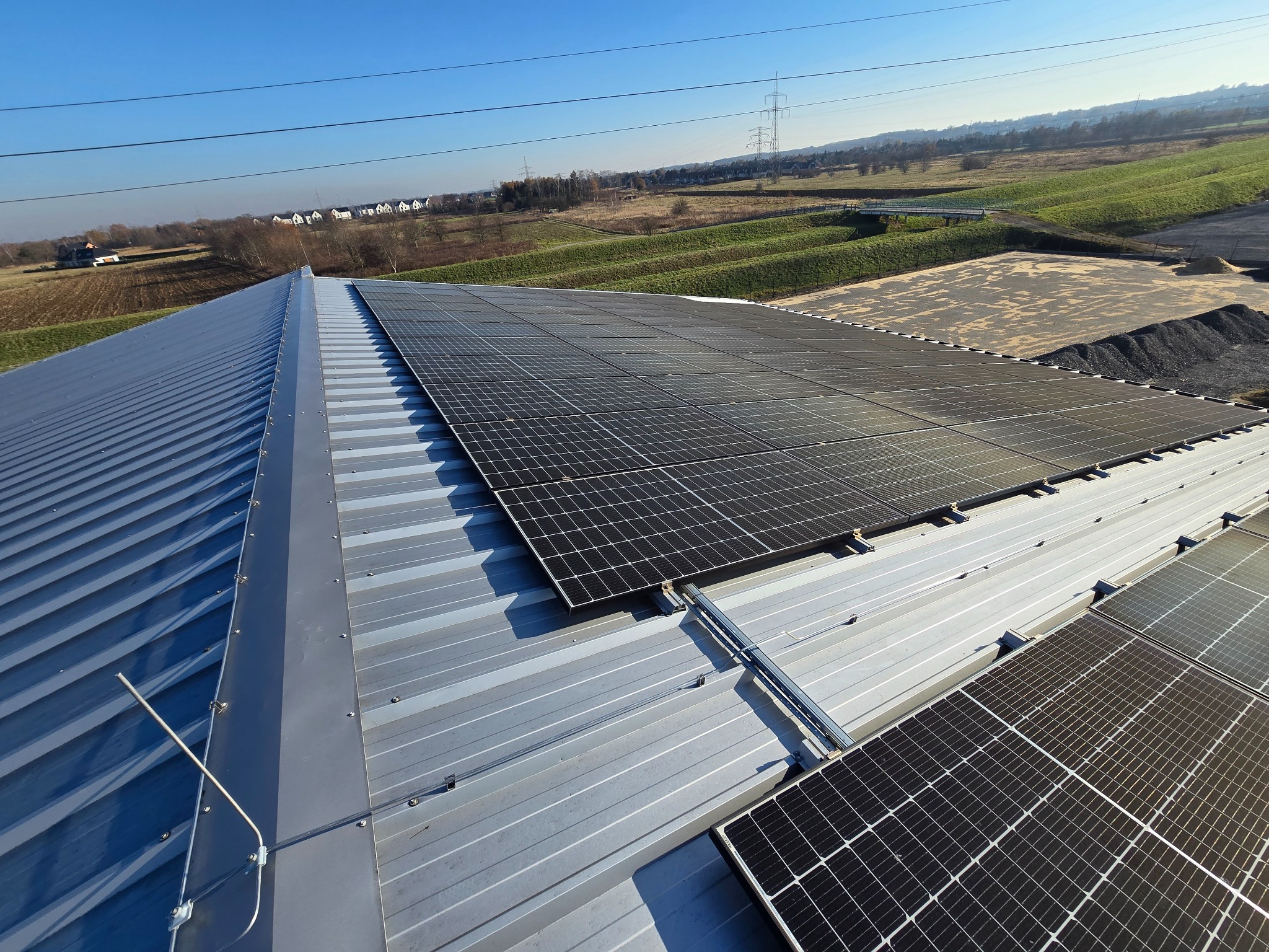 Panele fotowoltaiczne na metalowym dachu budynku, widok z góry, w tle krajobraz z liniami wysokiego napięcia i zabudowaniami.