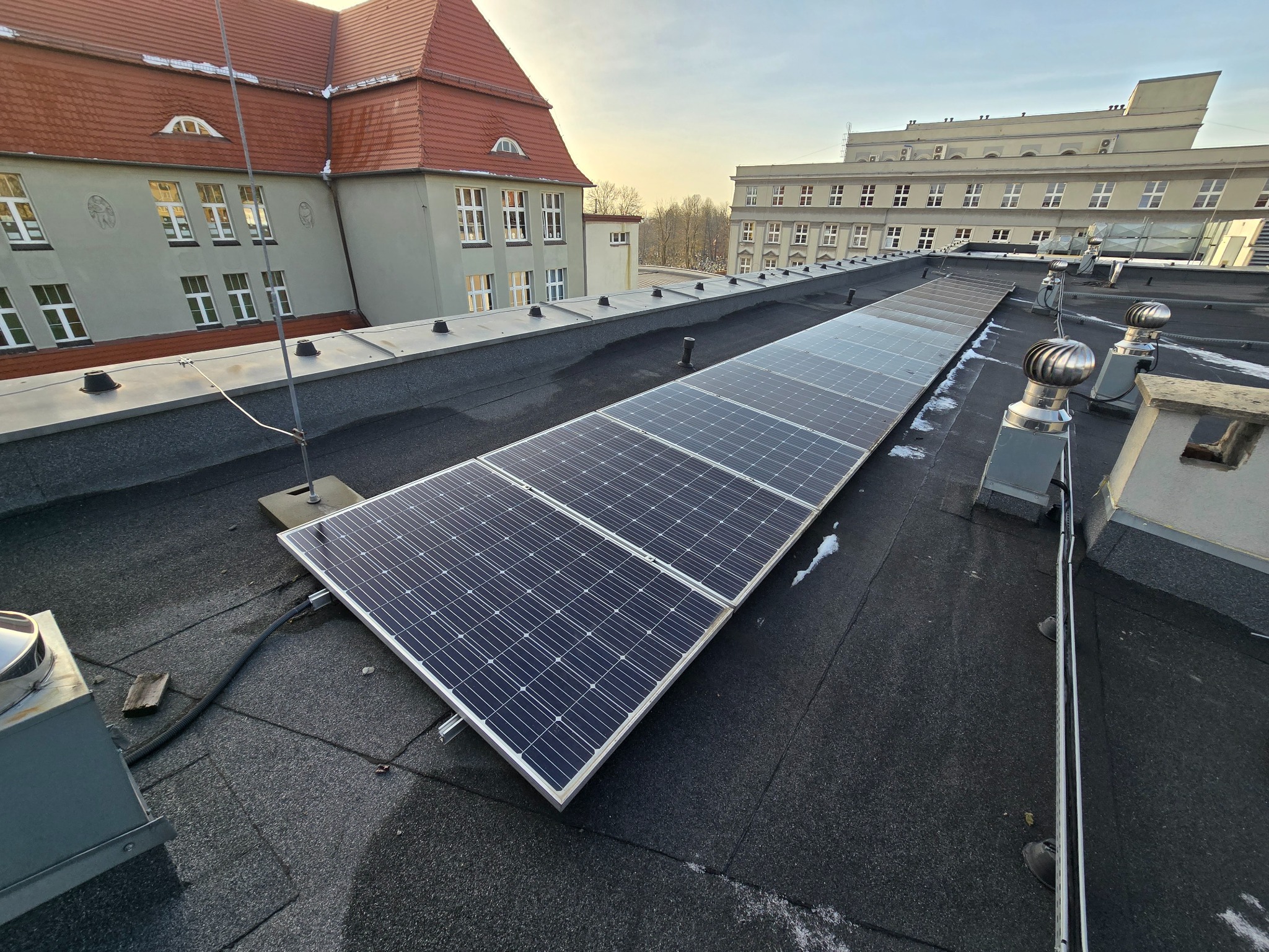 Rząd paneli fotowoltaicznych na płaskim dachu budynku, widoczne elementy montażowe i przewody, w tle budynki z ceglanymi dachami.