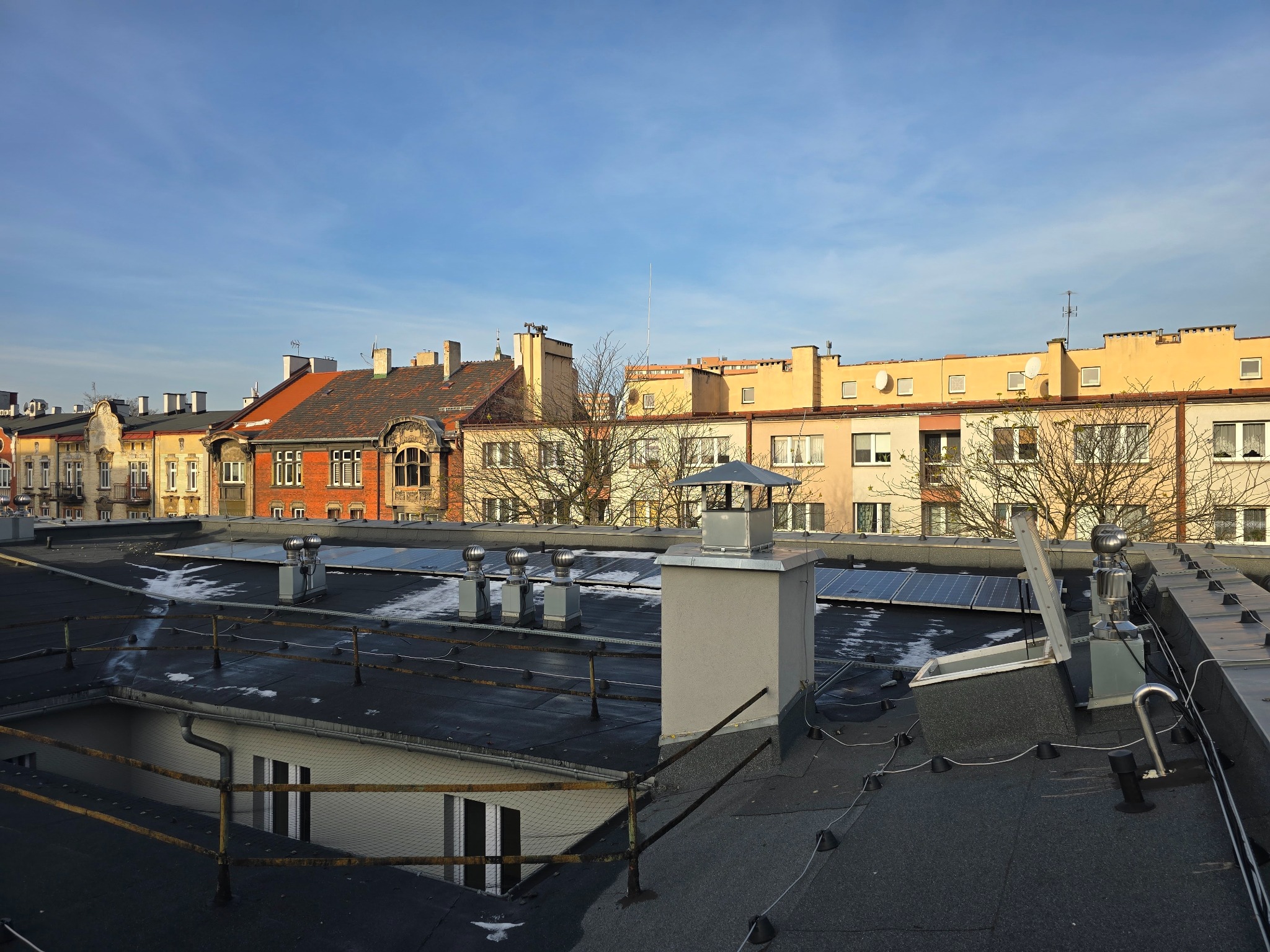 Panele fotowoltaiczne na dachu budynku w miejskiej zabudowie, widoczne kominy wentylacyjne i elementy instalacji odgromowej.