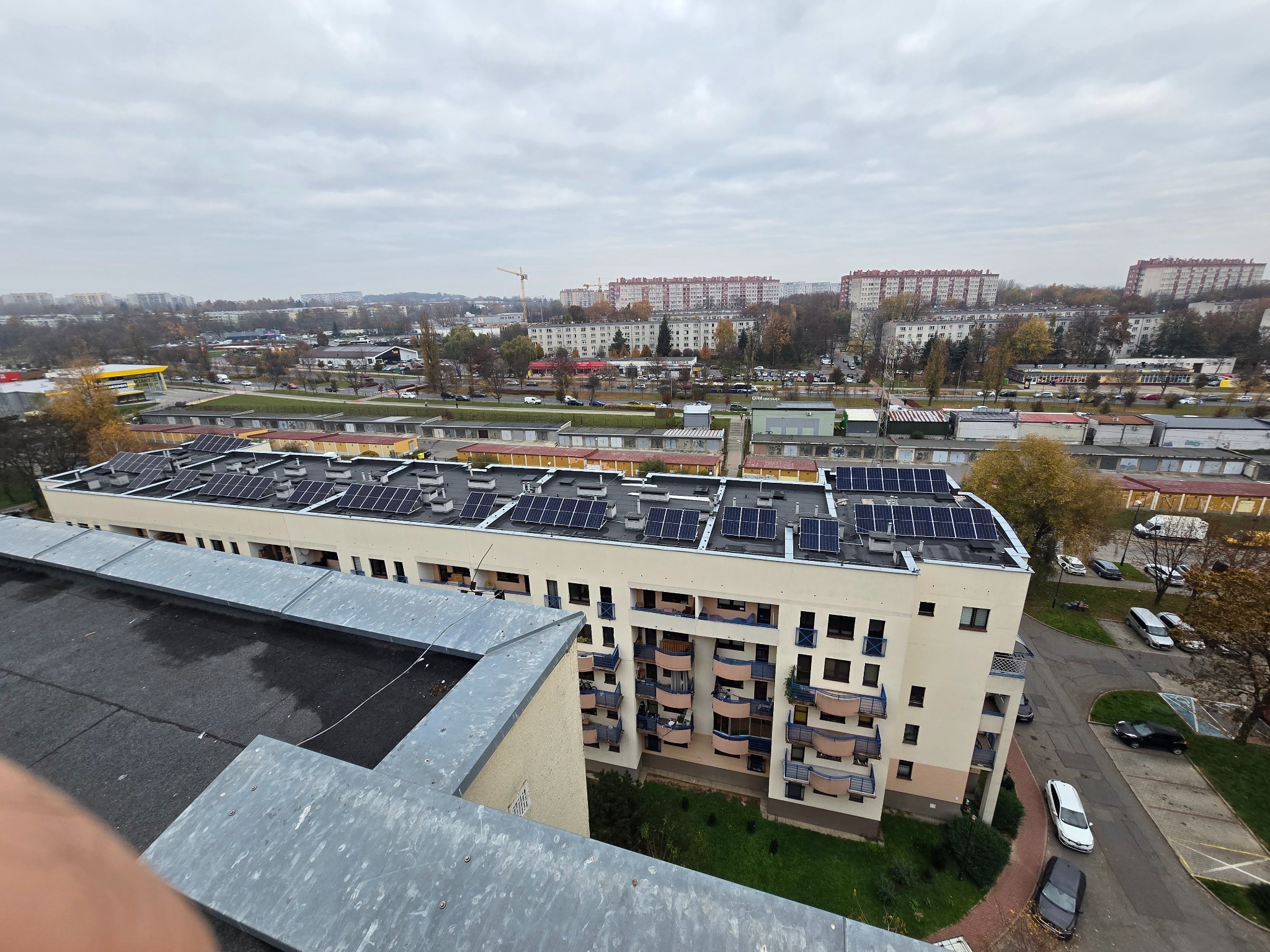 Widok z góry na budynek z panelami fotowoltaicznymi na dachu, w tle zabudowa miejska i pochmurne niebo.