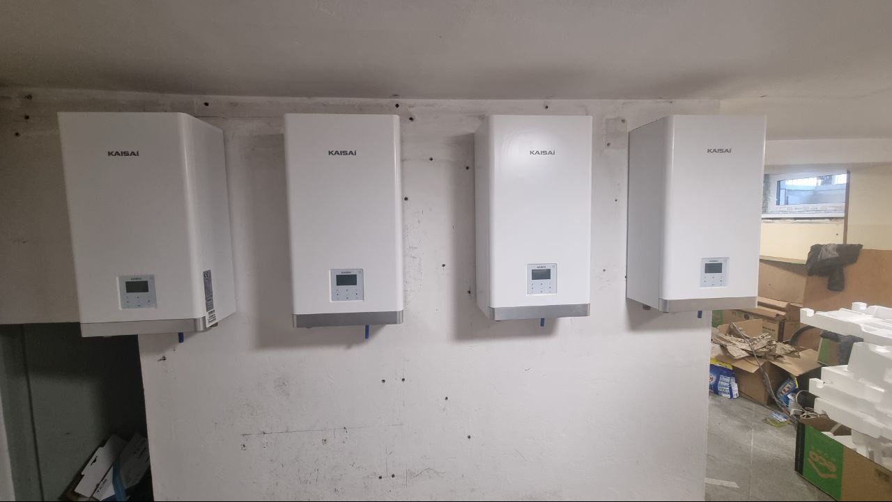 Cztery białe pompy ciepła marki Kaisai zamontowane na ścianie w pomieszczeniu gospodarczym, widoczne ślady po wierceniu i kartony w tle.