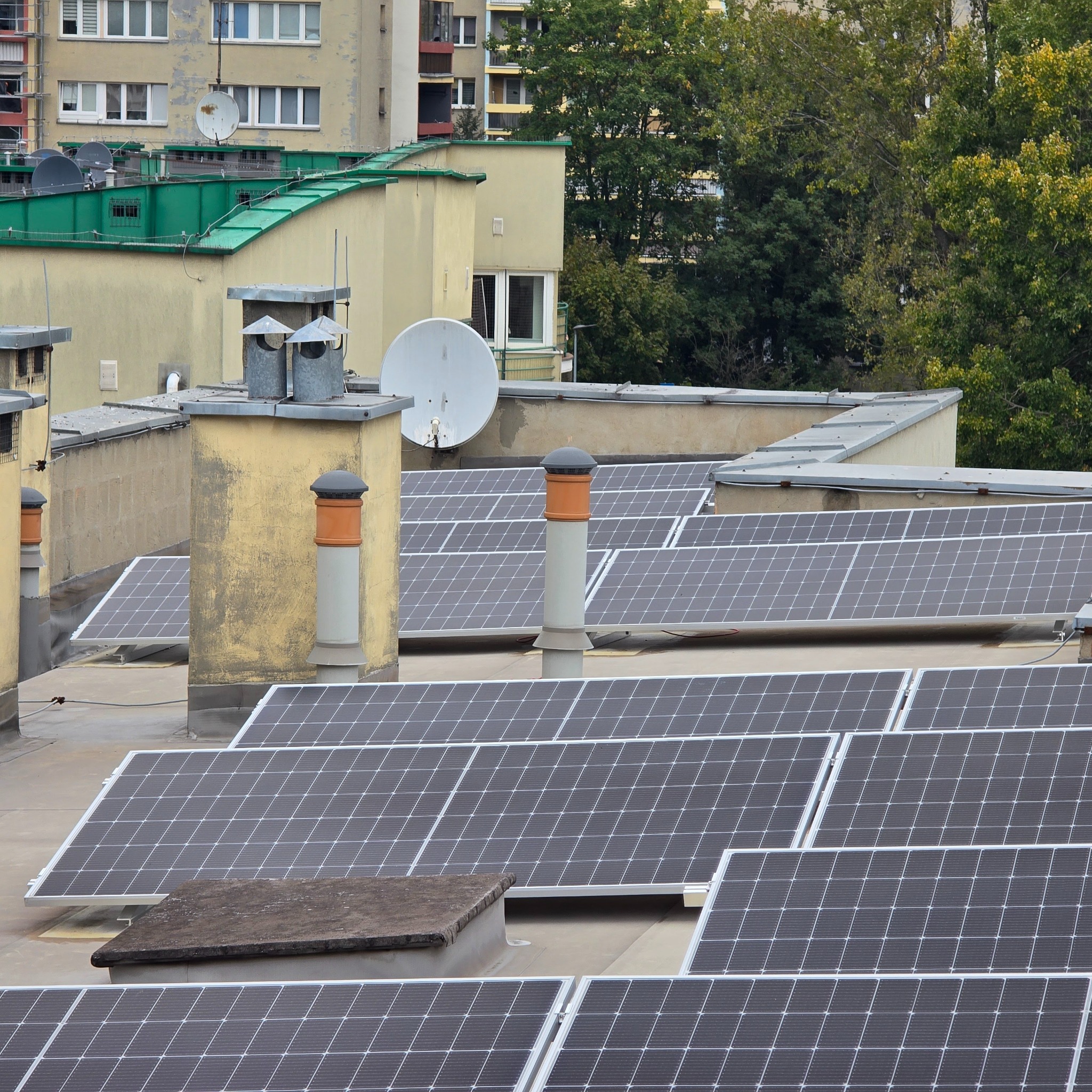 Panele fotowoltaiczne na płaskim dachu budynku mieszkalnego, widoczne kominy wentylacyjne i anteny satelitarne, w tle drzewa i inne budynki.
