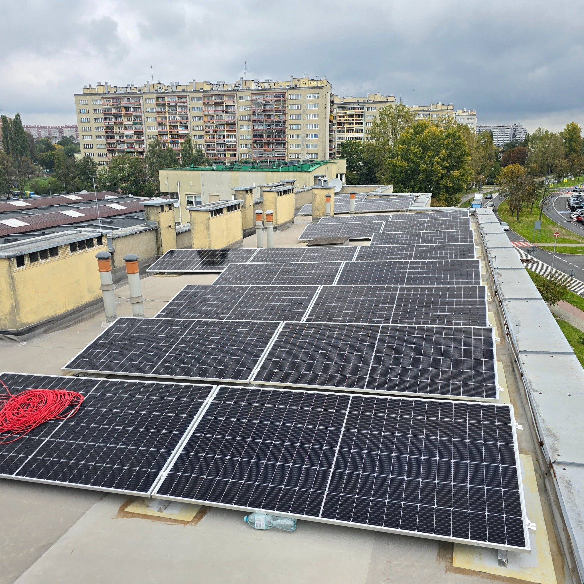 Widok z góry na panele fotowoltaiczne zamontowane na płaskim dachu budynku przemysłowego, z czerwonym kablem zwiniętym na jednym z paneli i butelką wody pod innym, w tle bloki mieszkalne.