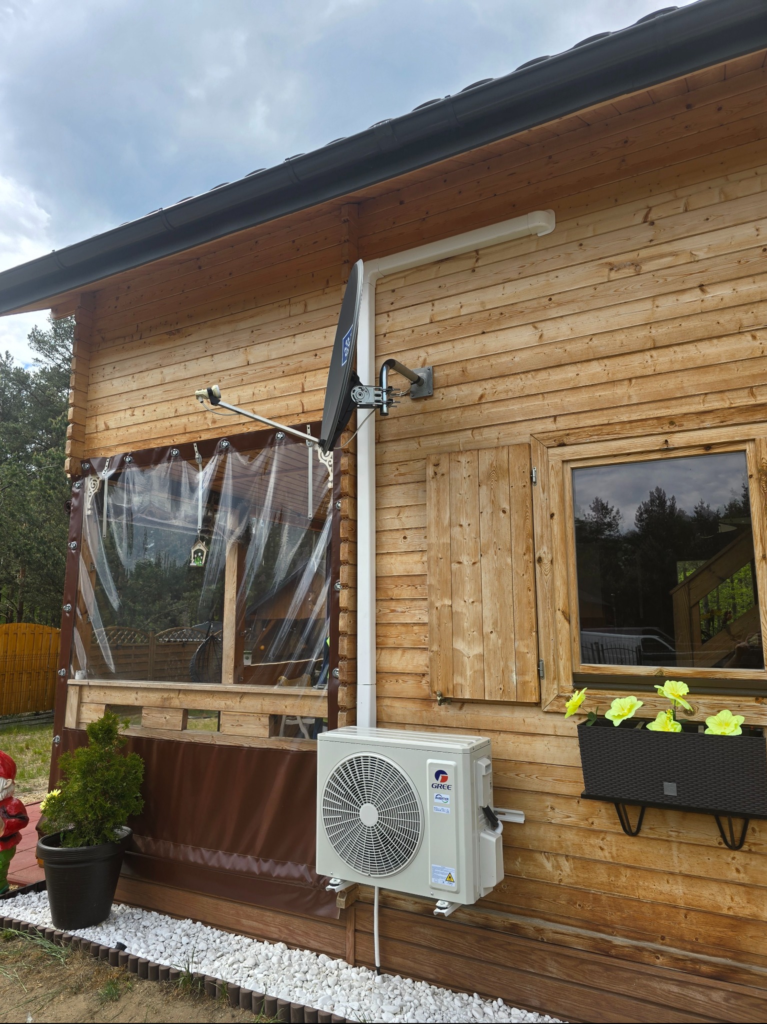 Biała klimatyzacja na drewnianej ścianie domku letniskowego z widoczną instalacją. Na ścianie zamontowana antena satelitarna.