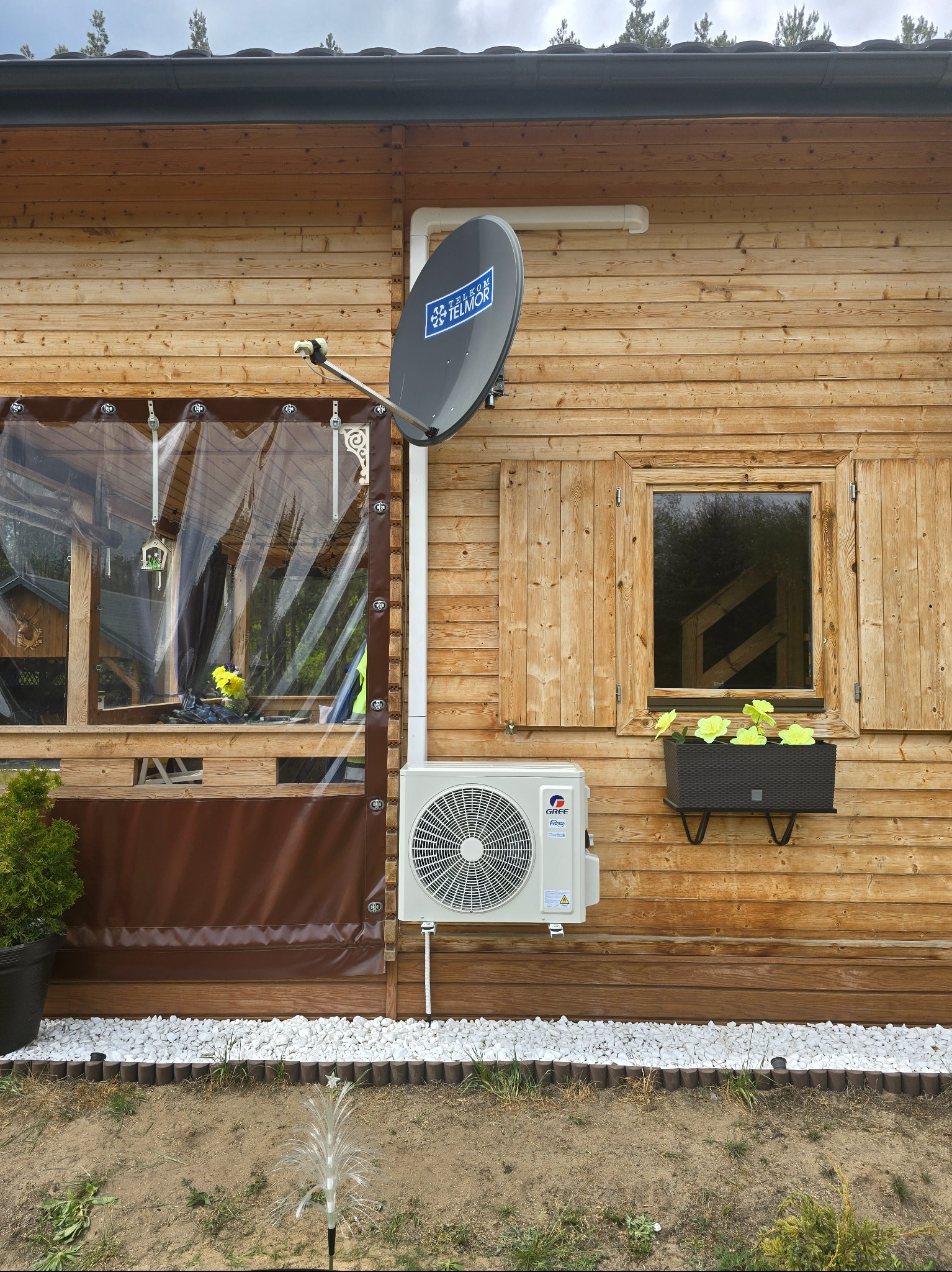 Klimatyzator GREE zamontowany na drewnianej elewacji domu z dekoracyjną anteną satelitarną i ozdobnym oświetleniem ogrodu. Widoczny fragment okna z roletami.