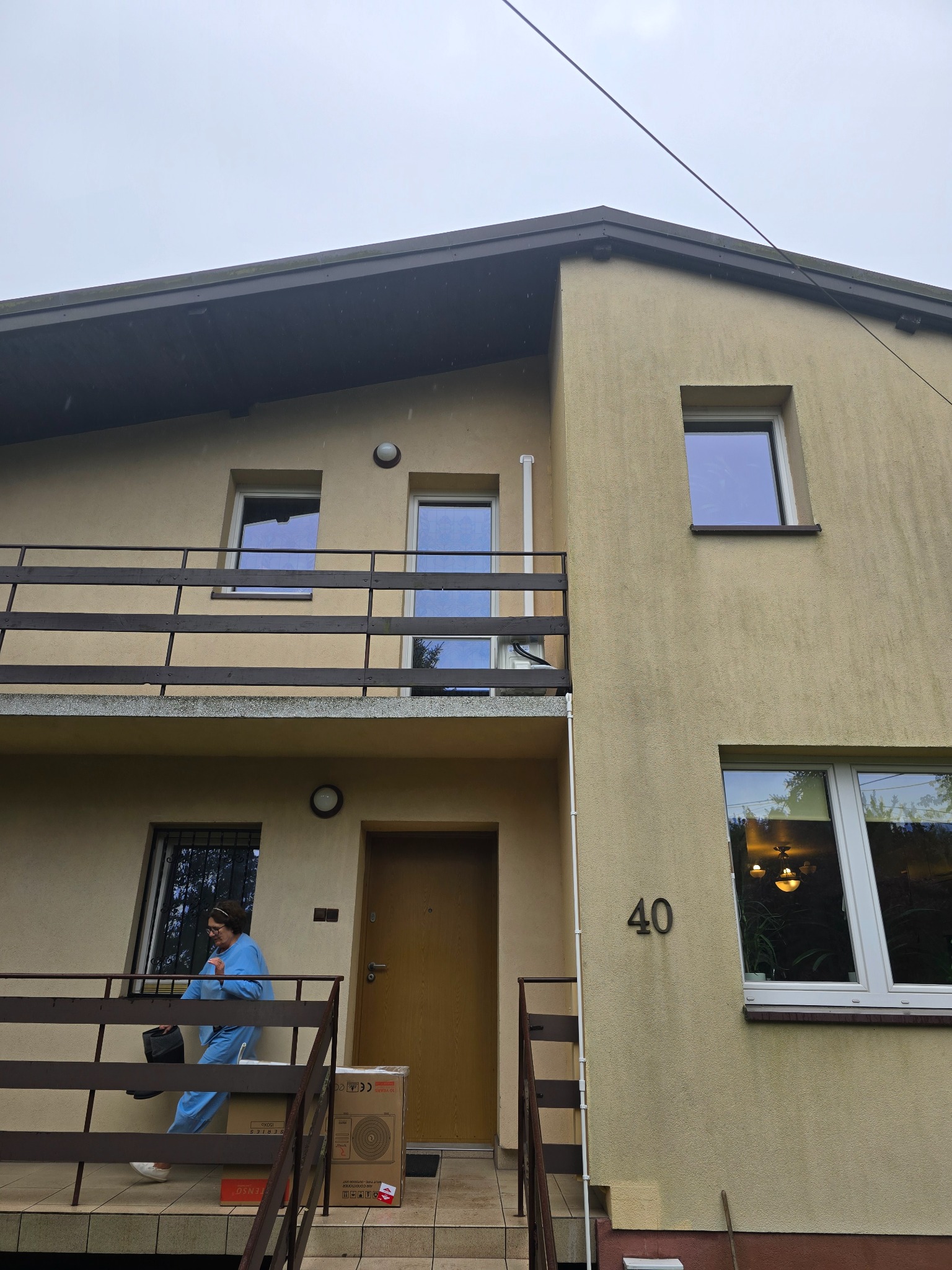 Dostawa klimatyzacji do domu: kobieta w niebieskim kombinezonie wnosi karton z klimatyzatorem po schodach do budynku z numerem 40.