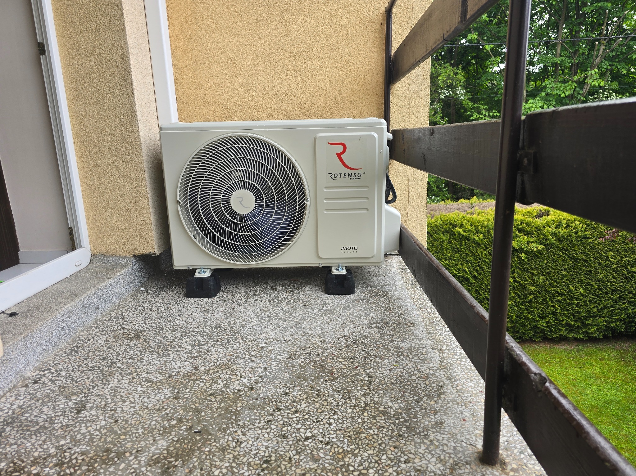 Klimatyzator Rotenso IMOTO zainstalowany na balkonie, widoczny z boku. Urządzenie stoi na gumowych podkładkach antywibracyjnych, tło stanowi żółta ściana budynku i zielony żywopłot.