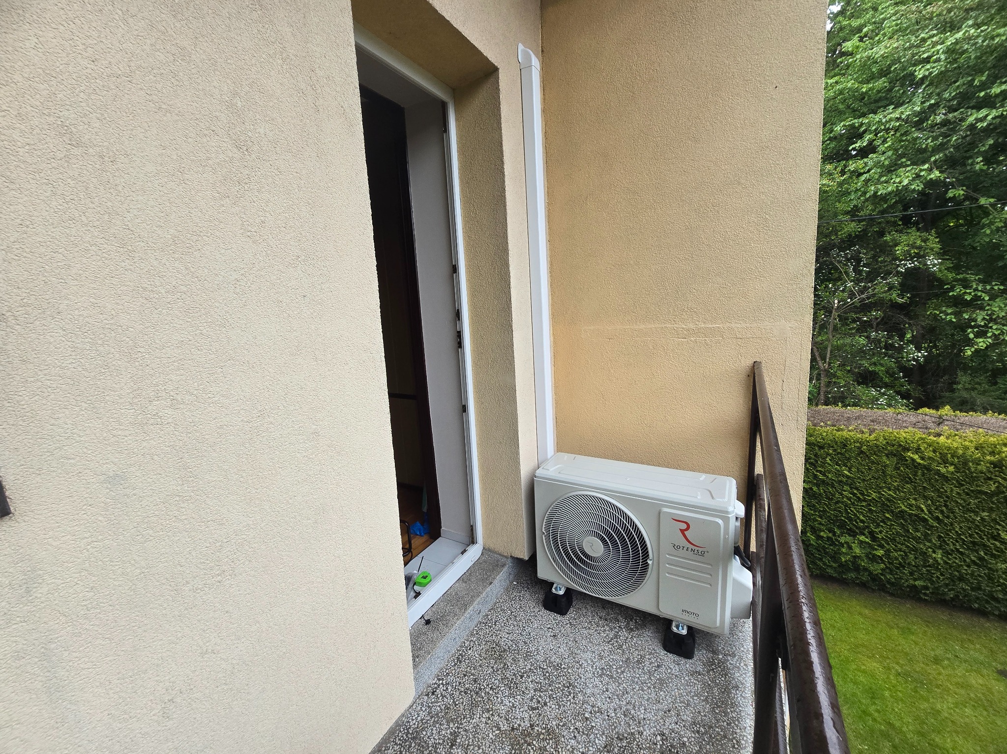 Biały klimatyzator Rotenso IMOTO na balkonie, obok drzwi balkonowych. Widoczna fasada budynku i zieleń w tle. Elementy instalacji klimatyzacji.