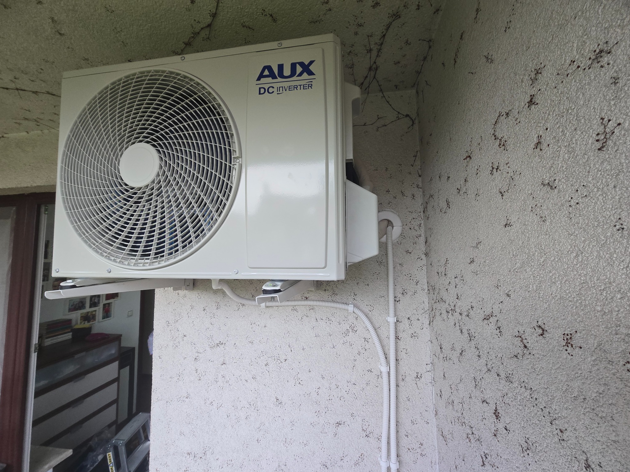 Biała klimatyzacja AUX DC Inverter zamontowana na elewacji budynku z widocznymi przewodami. Widok z dołu, z fragmentem wnętrza w tle.