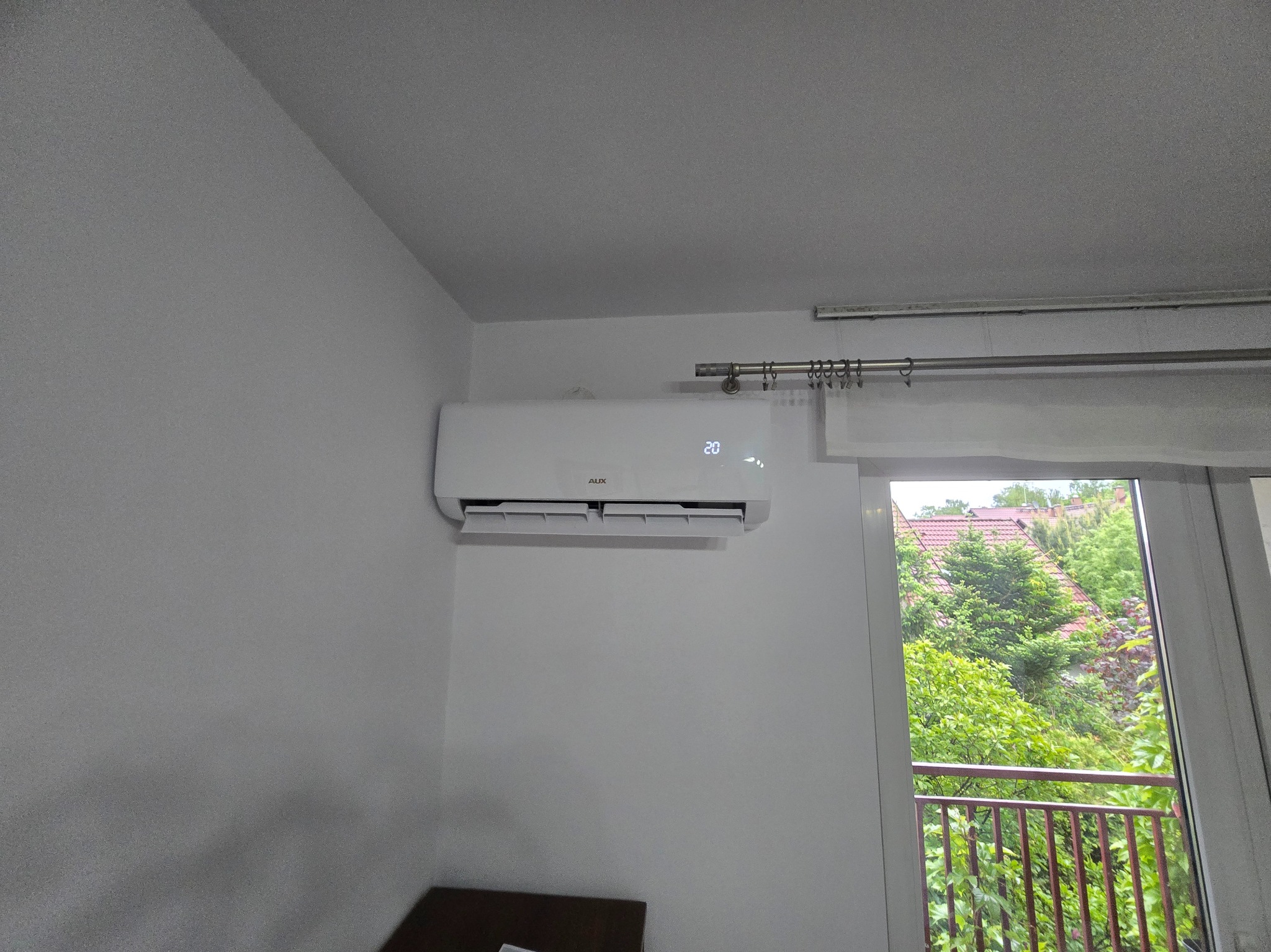 Biały klimatyzator AUX na ścianie w jasnym pokoju, widok z balkonu z zielenią w tle. Na wyświetlaczu urządzenia widoczna temperatura 20 stopni.