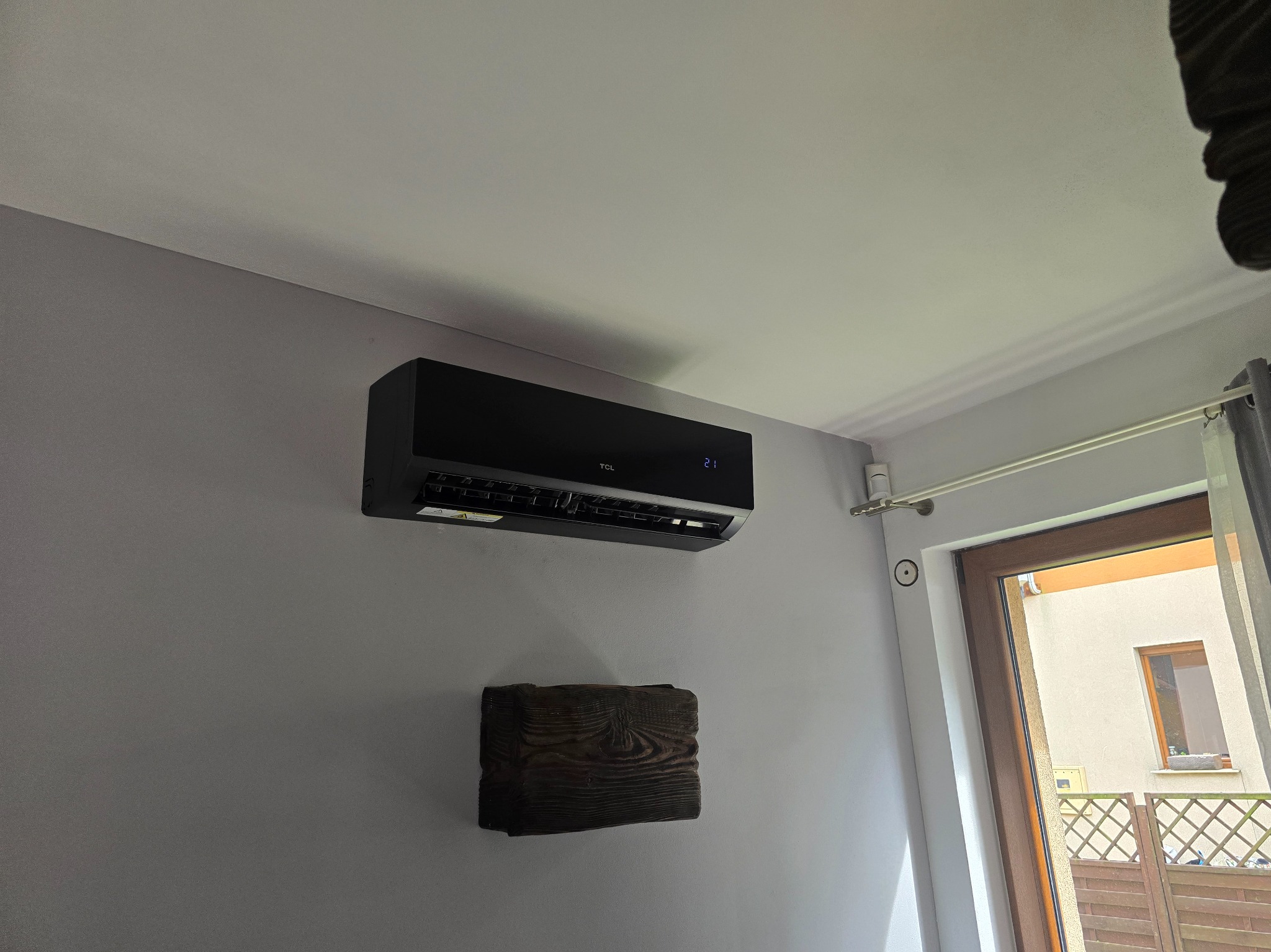 Czarna klimatyzacja TCL na ścianie w mieszkaniu w Krakowie. Widoczny drewniany element dekoracyjny pod klimatyzatorem i okno z widokiem na zewnątrz.