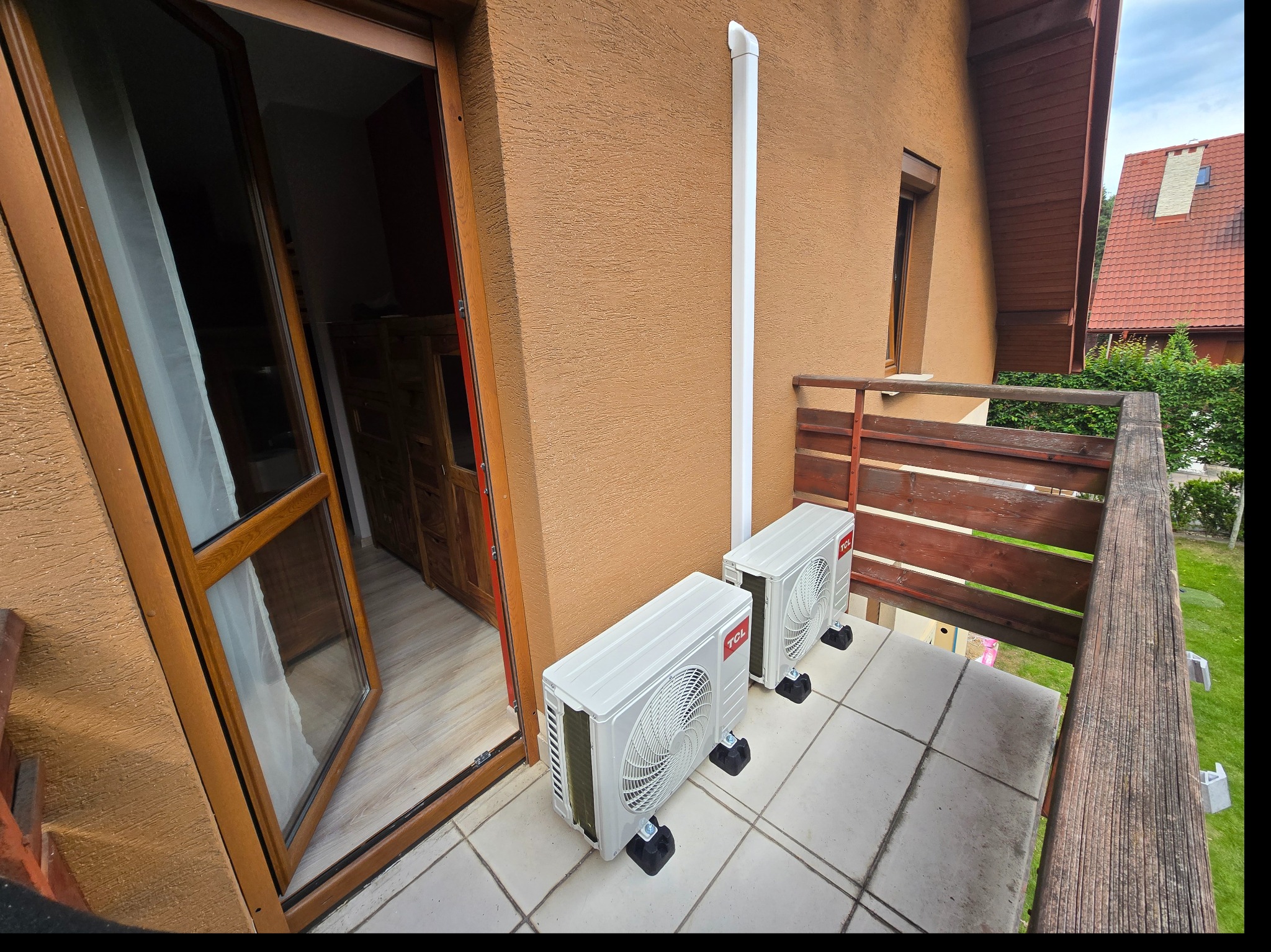 Dwa białe klimatyzatory TCL Elite na balkonie z brązową balustradą, widoczne z zewnątrz. Rura odprowadzająca na elewacji budynku. Otwarte drzwi balkonowe.