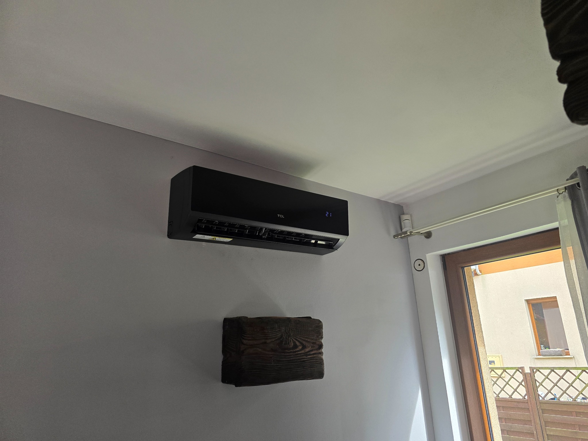 Czarna klimatyzacja TCL zamontowana na szarej ścianie w pokoju. Pod nią drewniana ozdoba. Widoczne okno z zasłoną i fragment balkonu.