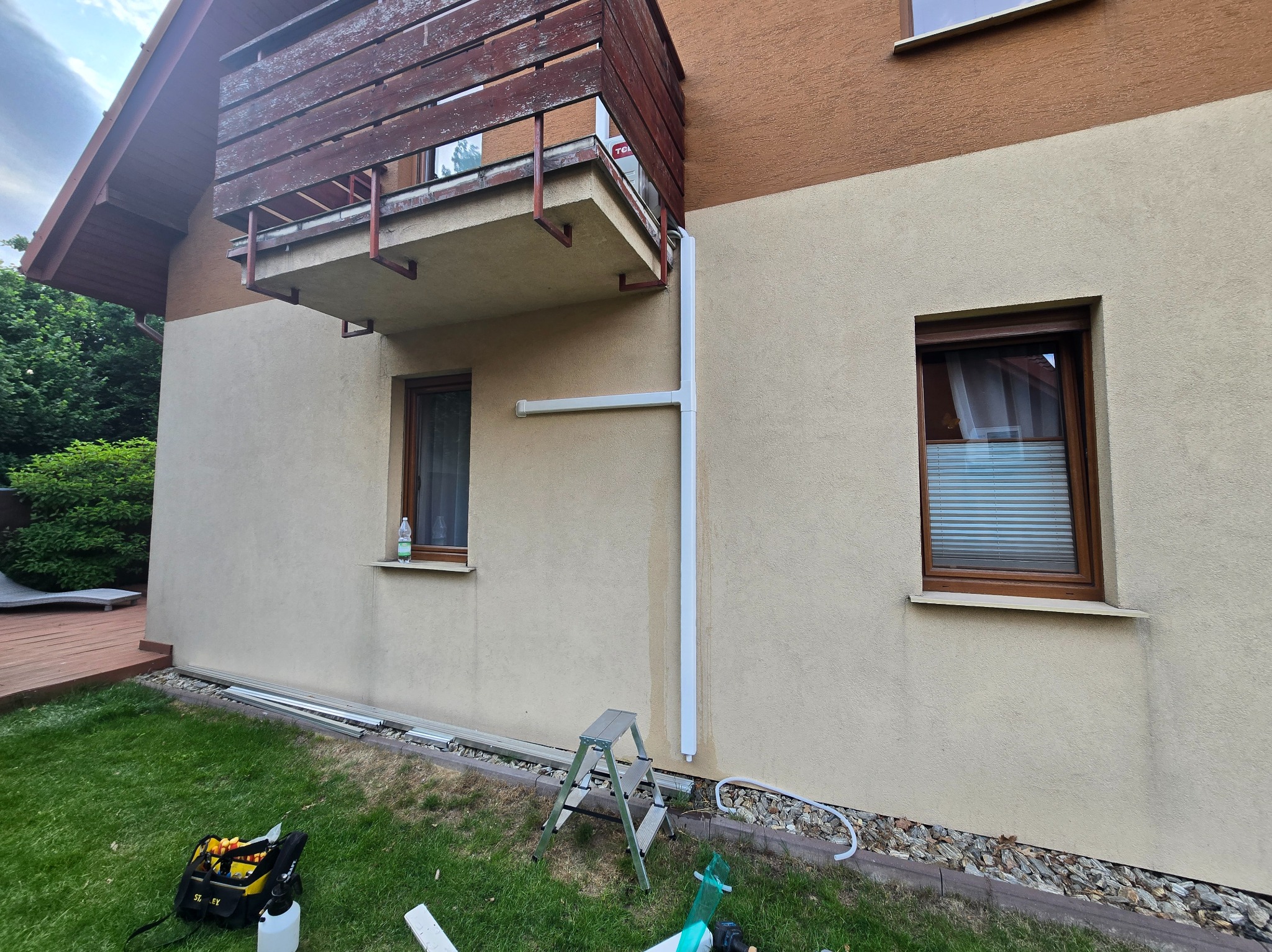 Elewacja domu z nową instalacją klimatyzacji: rury prowadzone wzdłuż ściany, drabina i narzędzia na trawniku. Widoczny balkon z brązową balustradą.