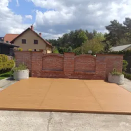 Nowoczesny taras z desek kompozytowych w odcieniach brązu, otoczony ceglanym murem i zielenią ogrodu. Słoneczny dzień, błękitne niebo z chmurami w tle.