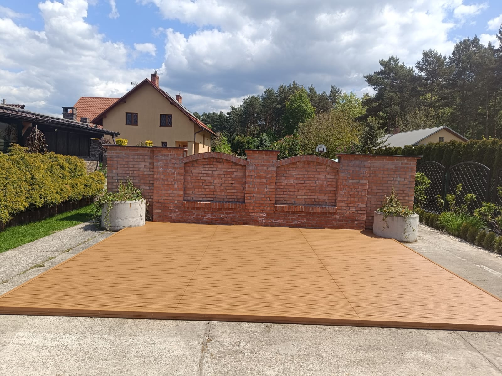 Nowoczesny taras z desek kompozytowych w odcieniach brązu, otoczony ceglanym murem i zielenią ogrodu. Słoneczny dzień, błękitne niebo z chmurami w tle.