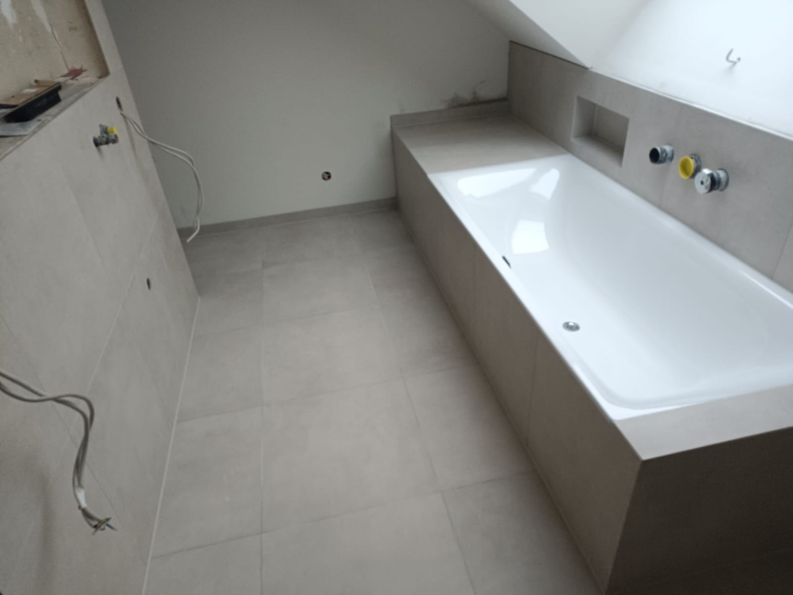 Wykończona łazienka na poddaszu ze skośnym sufitem. Biała wanna obudowana płytkami, szare płytki na podłodze i ścianach. Widoczne instalacje wodne i elektryczne.