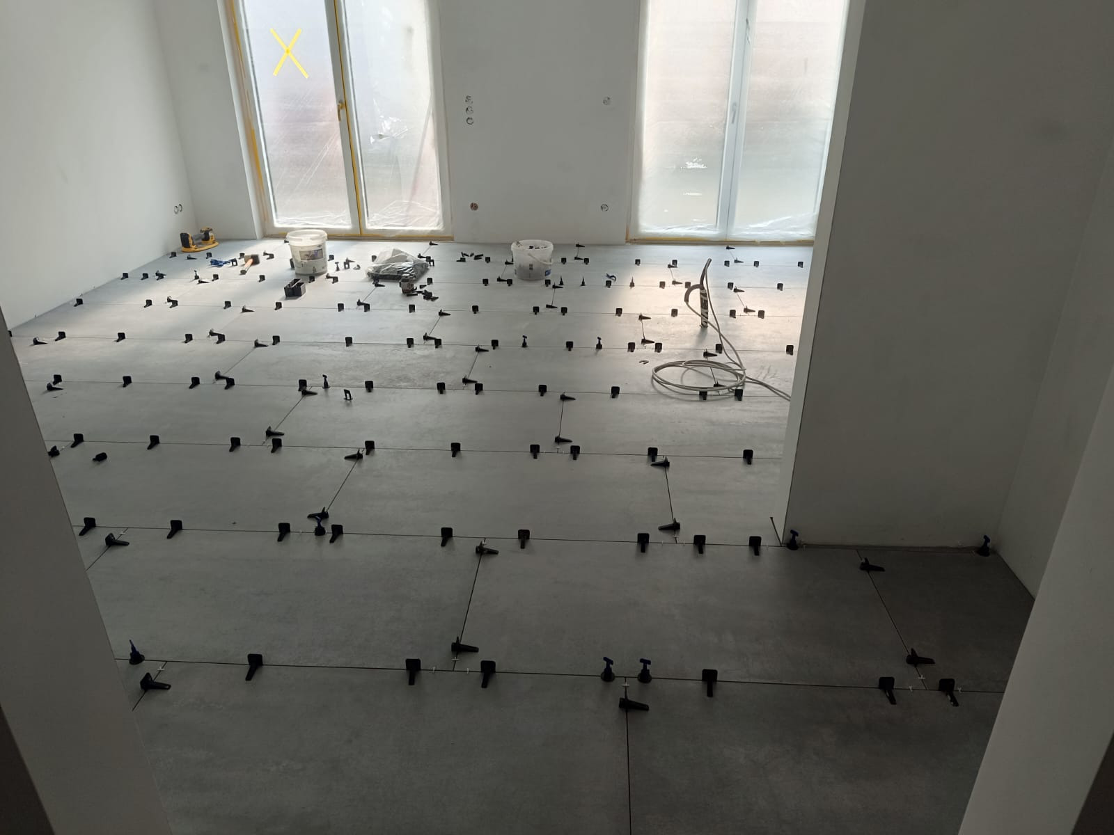 Układanie szarych płytek podłogowych z użyciem systemu poziomowania. Widoczne narzędzia, wiaderka i okna zabezpieczone folią malarską. Etap prac wykończeniowych.