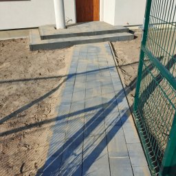 USŁUGI BESZTERDA JAKUB - Świeżo ułożona ścieżka z szarej kostki brukowej prowadzi od zielonej bramki ogrodzeniowej do wejścia domu z kolumną i drewnianymi drzwiami, teren wokół wysypany piaskiem.