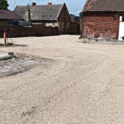 USŁUGI BESZTERDA JAKUB - Szeroki plac pokryty drobnym kruszywem, otoczony starymi budynkami z cegły i dachówką, widoczne ślady wcześniejszych prac ziemnych.