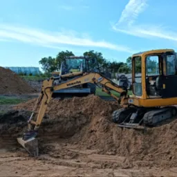 Żółta mini koparka Komatsu podczas prac ziemnych, wykop pod fundament, w tle druga maszyna i pryzma piasku.
