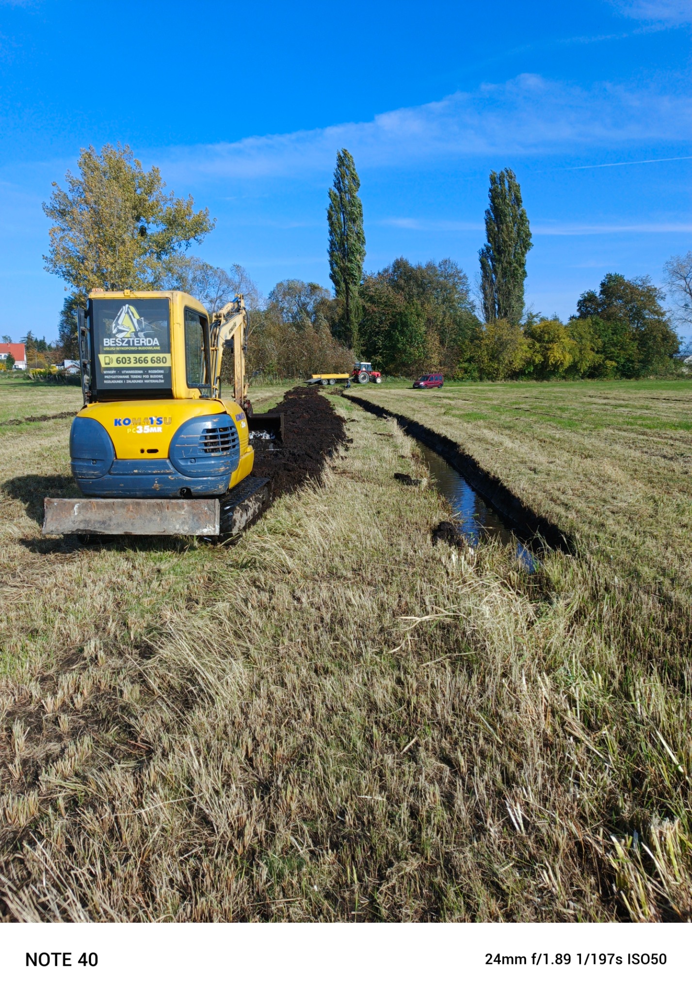 Żółto-niebieska mini koparka Komatsu z łyżką, wykonująca wykop w zarośniętym polu, w tle widoczny traktor z przyczepą i czerwony samochód osobowy, błękitne niebo z lekkimi chmurami.