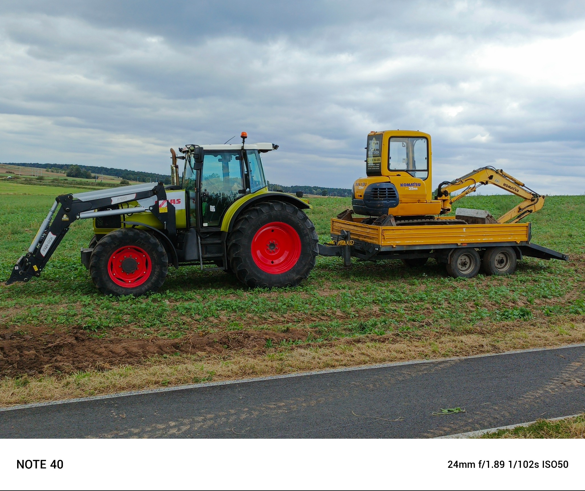 Żółty minikoparka Komatsu PC35MR na przyczepie, ciągnięta przez zielony ciągnik rolniczy Claas z czerwonymi felgami, na tle pola i pochmurnego nieba.