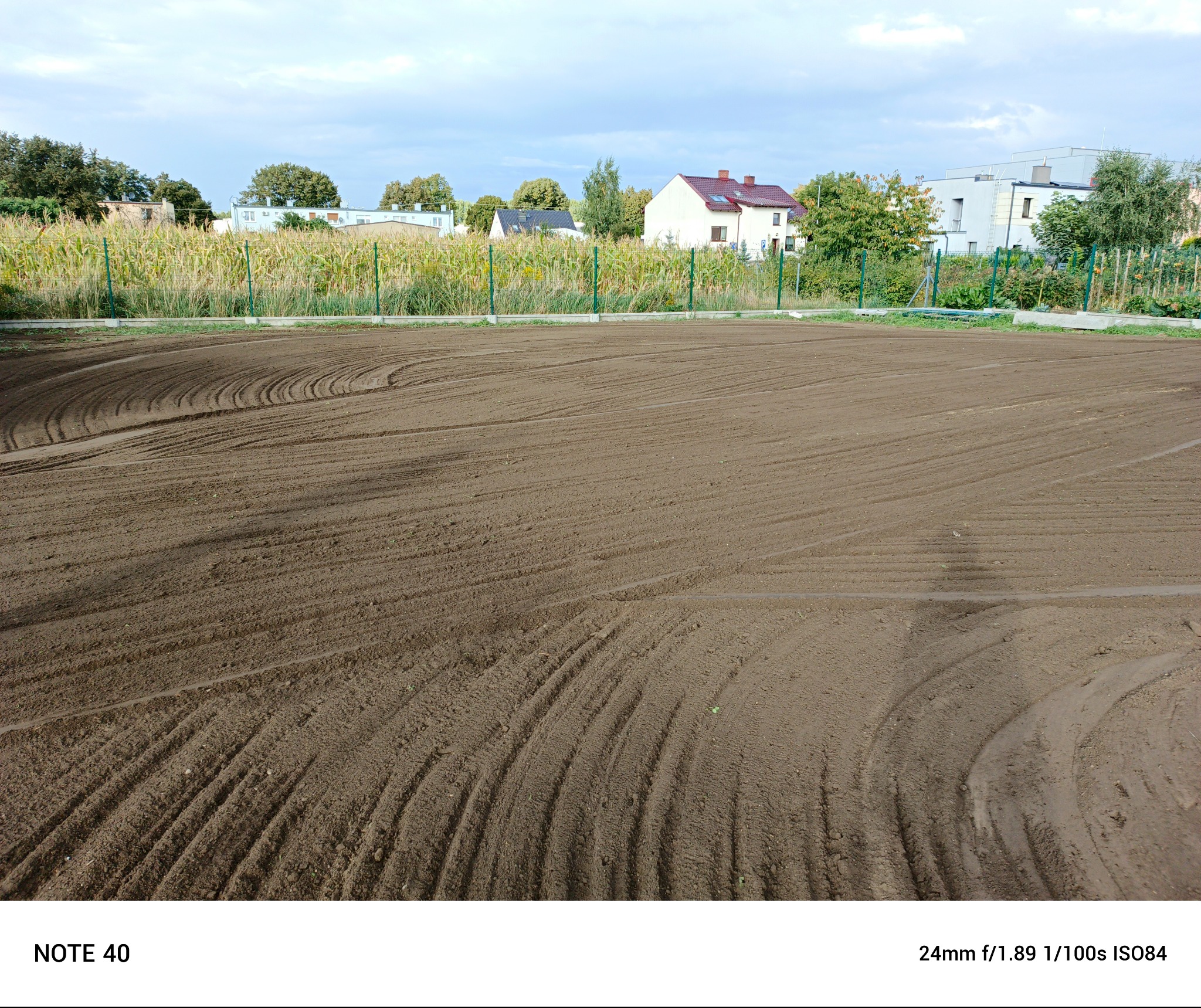 Przygotowany, wyrównany teren pod przyszłą inwestycję, widoczne ślady po maszynach rolniczych, w tle budynki mieszkalne i zielone ogrodzenie.