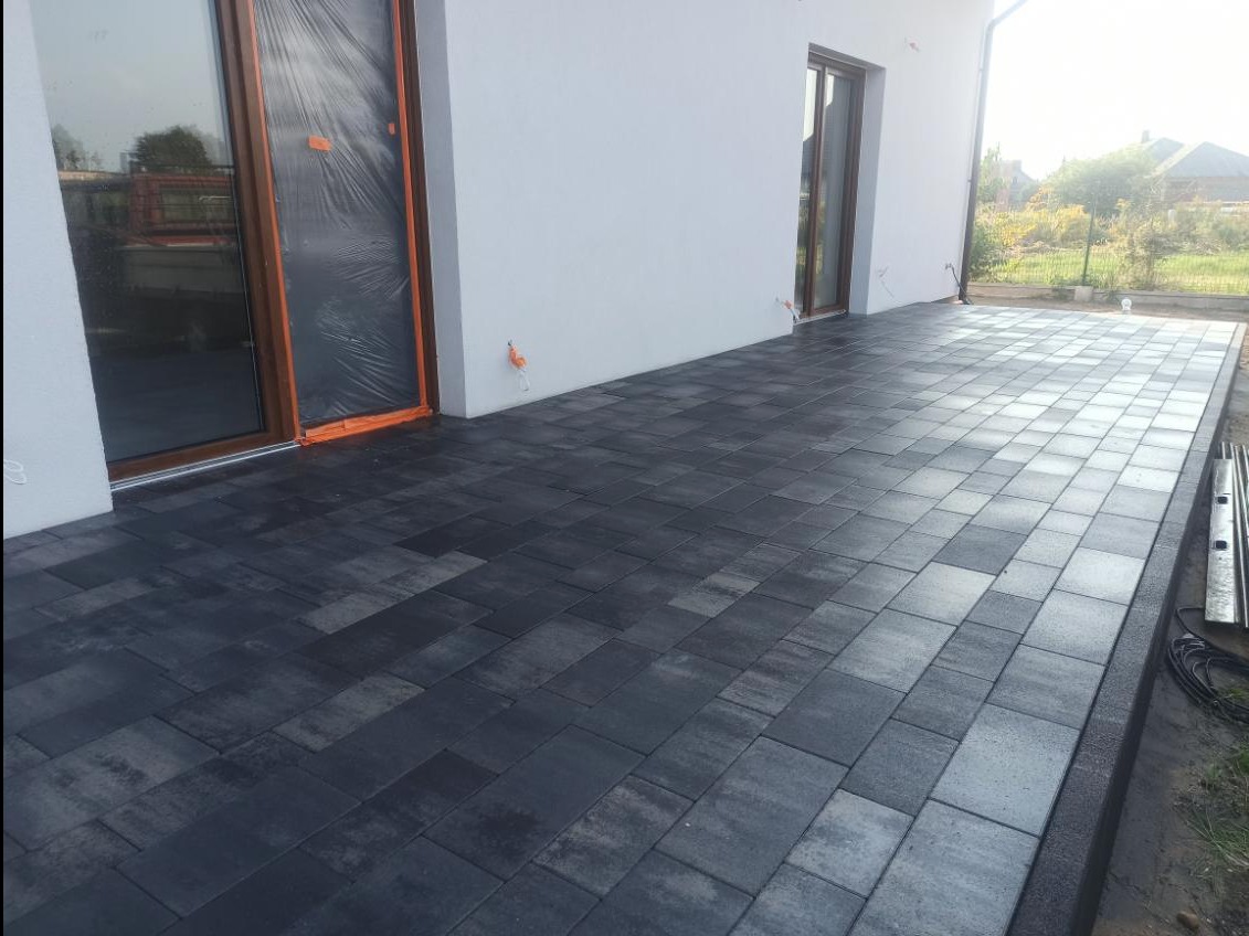 Nowoczesny taras z ciemnej kostki brukowej ułożonej przy białym budynku z dużymi oknami, jedno z nich zabezpieczone folią ochronną.