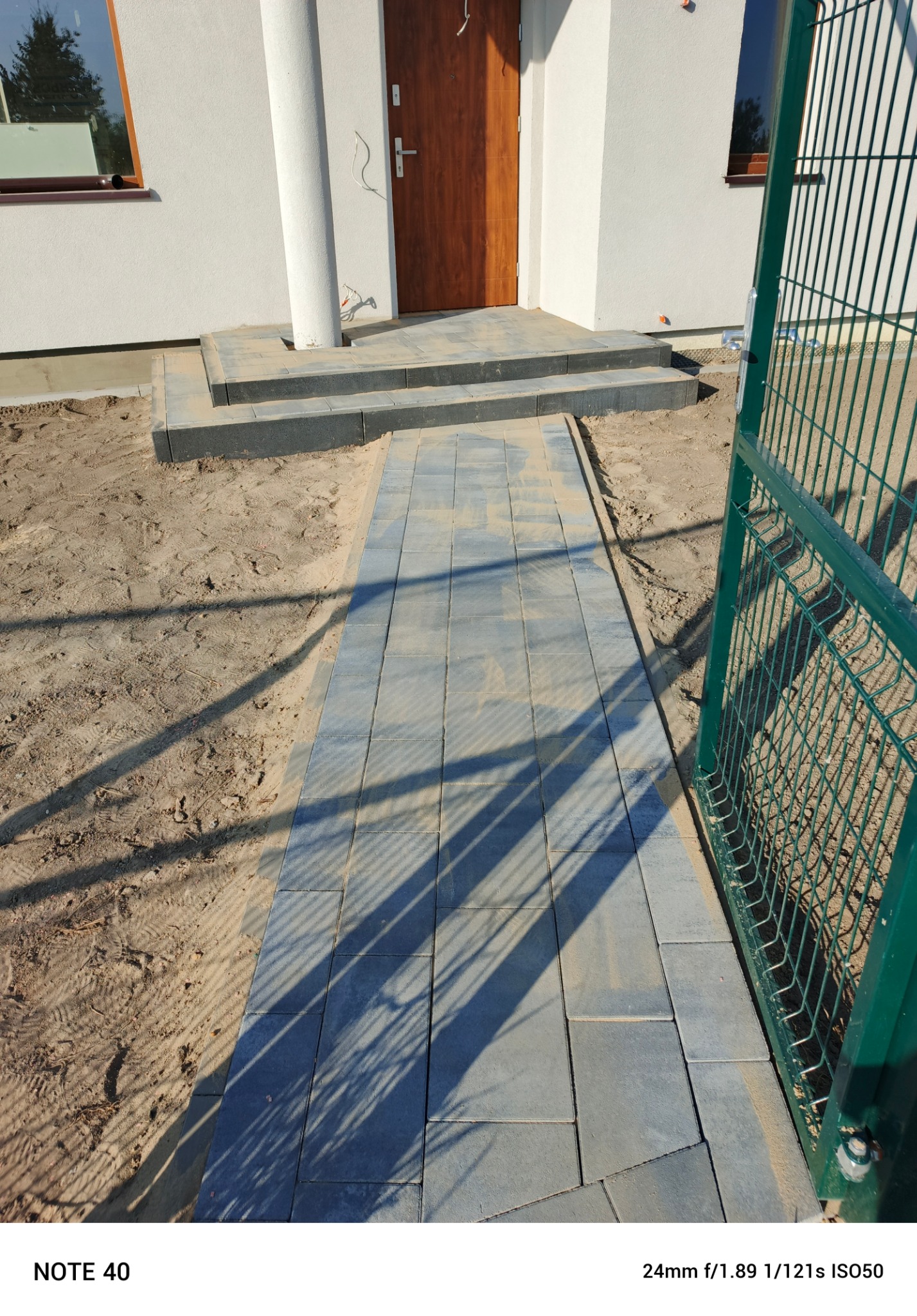 Świeżo ułożona ścieżka z szarej kostki brukowej prowadzi od zielonej bramki ogrodzeniowej do wejścia domu z kolumną i drewnianymi drzwiami, teren wokół wysypany piaskiem.