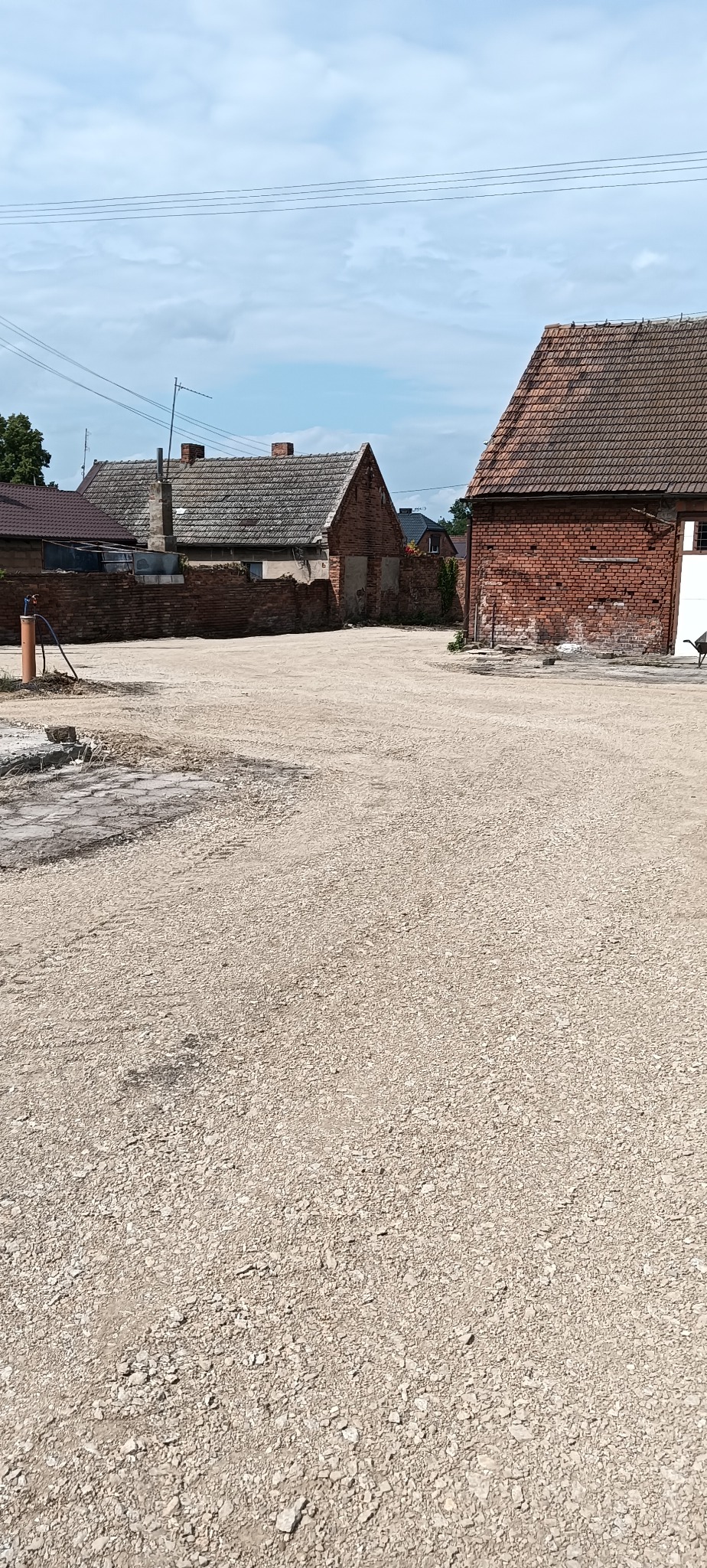 Szeroki plac pokryty drobnym kruszywem, otoczony starymi budynkami z cegły i dachówką, widoczne ślady wcześniejszych prac ziemnych.