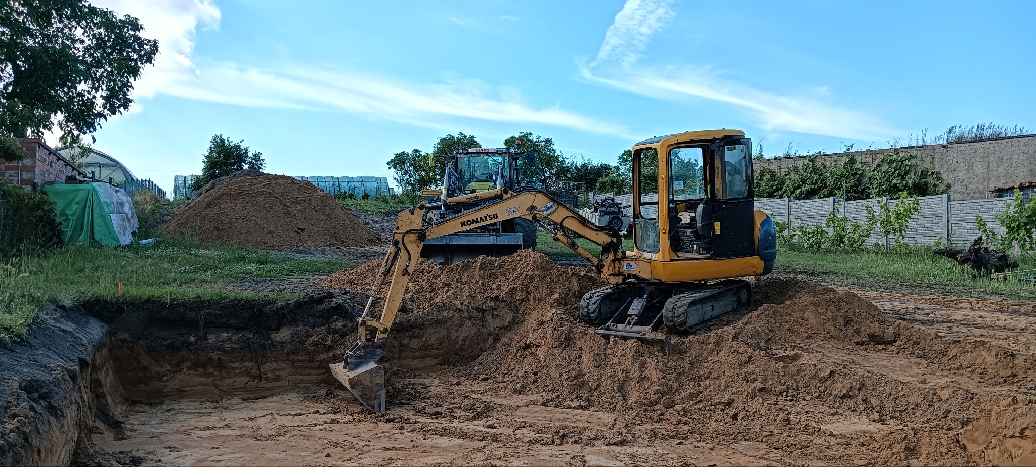 Żółta mini koparka Komatsu podczas prac ziemnych, wykop pod fundament, w tle druga maszyna i pryzma piasku.