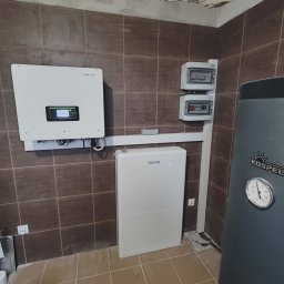 Instalacje Fotowoltaiczne ,,J&J'' Jakub Jankowiak - Biała bateria magazynu energii Sofar i falownik fotowoltaiczny tej samej marki zamontowane na brązowych płytkach ściennych w pomieszczeniu z wykończonym, ale niepomalowanym sufitem, obok szary...