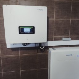 Instalacje Fotowoltaiczne ,,J&J'' Jakub Jankowiak - Biały inwerter i akumulator firmy SOFAR zamontowane na brązowej, kafelkowanej ścianie, obok liczników elektrycznych.