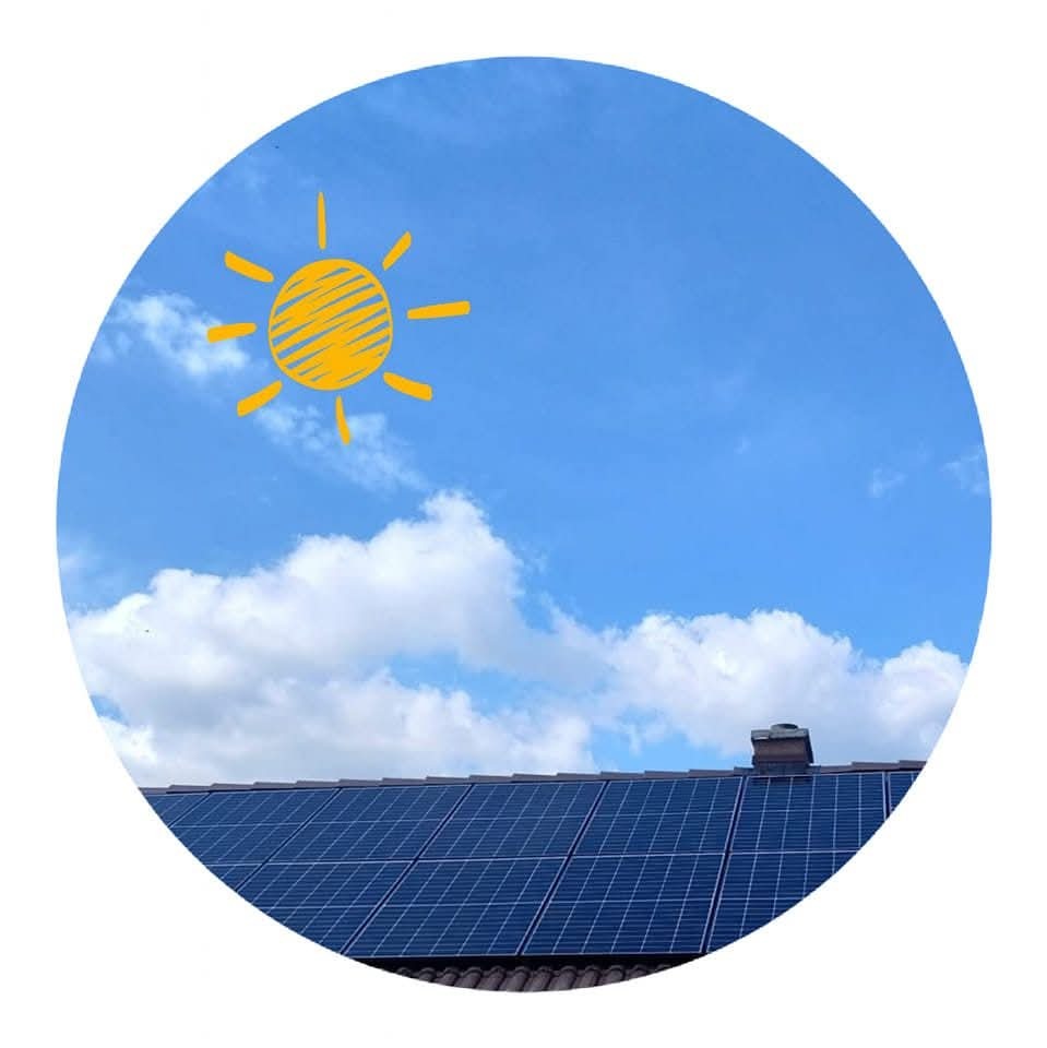 Niebieskie panele słoneczne na dachu domu, na tle błękitnego nieba z białymi obłokami i rysunkowym słońcem w rogu, ujęte w okrągłą ramę.