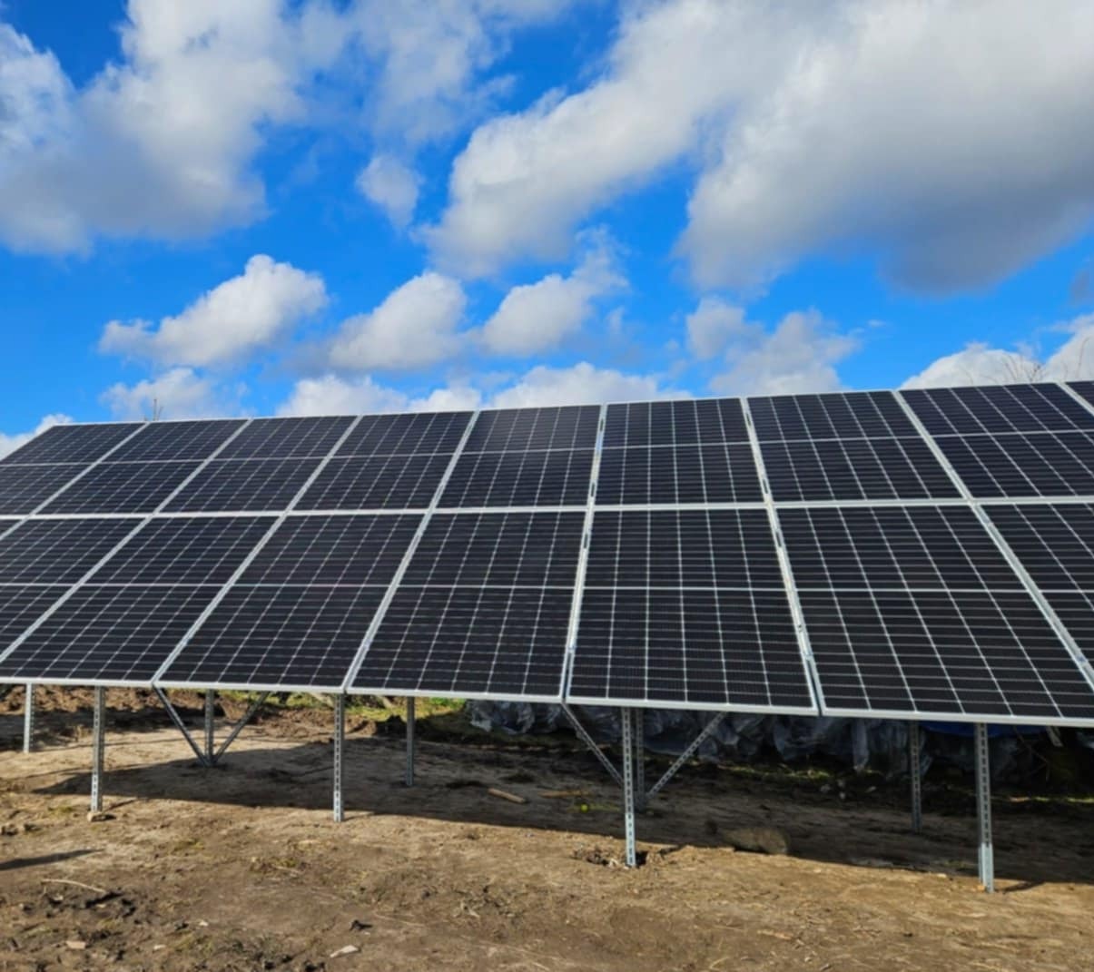 Rząd paneli fotowoltaicznych na metalowej konstrukcji, z widocznymi chmurami na niebie w tle, umieszczony na nierównym, ziemistym terenie.