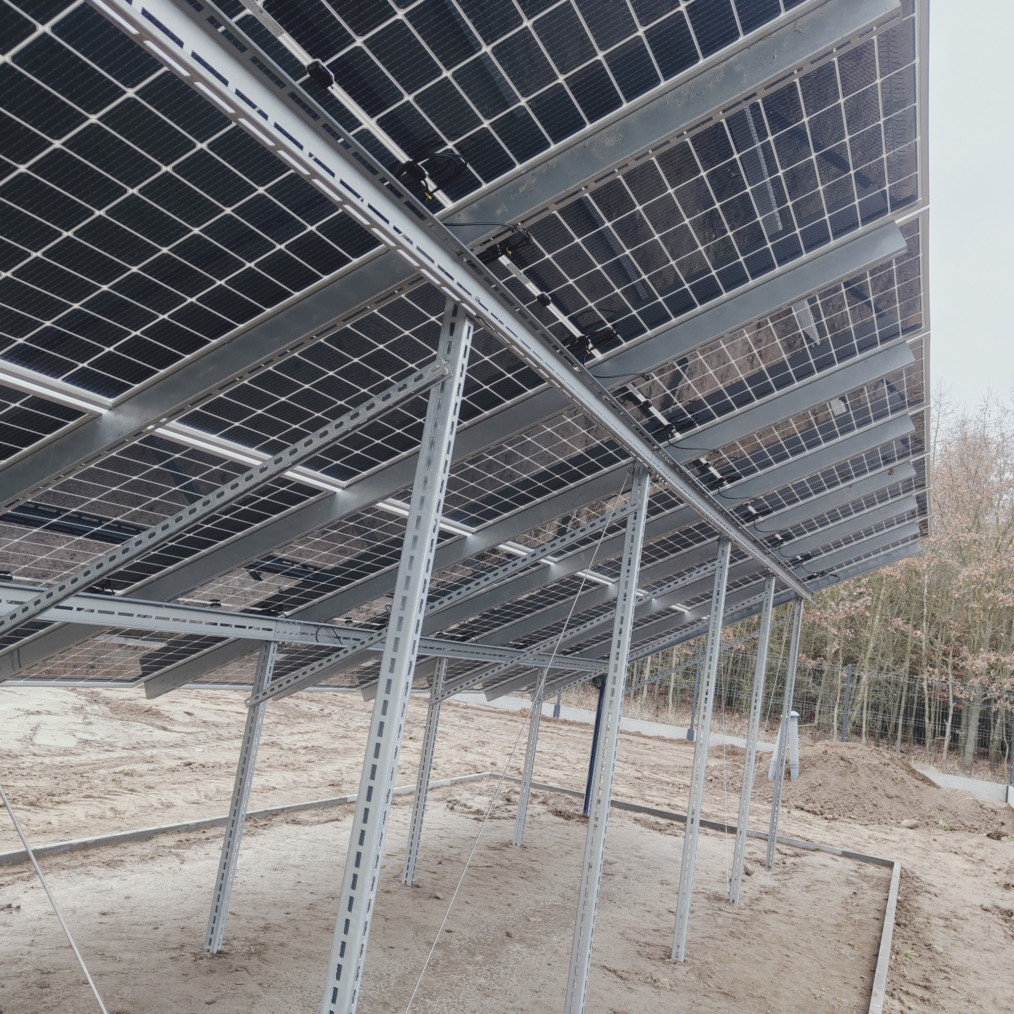 Konstrukcja wsporcza pod panele słoneczne z widocznymi elementami montażowymi i kablami, teren budowy z piaskiem i drzewami w tle.