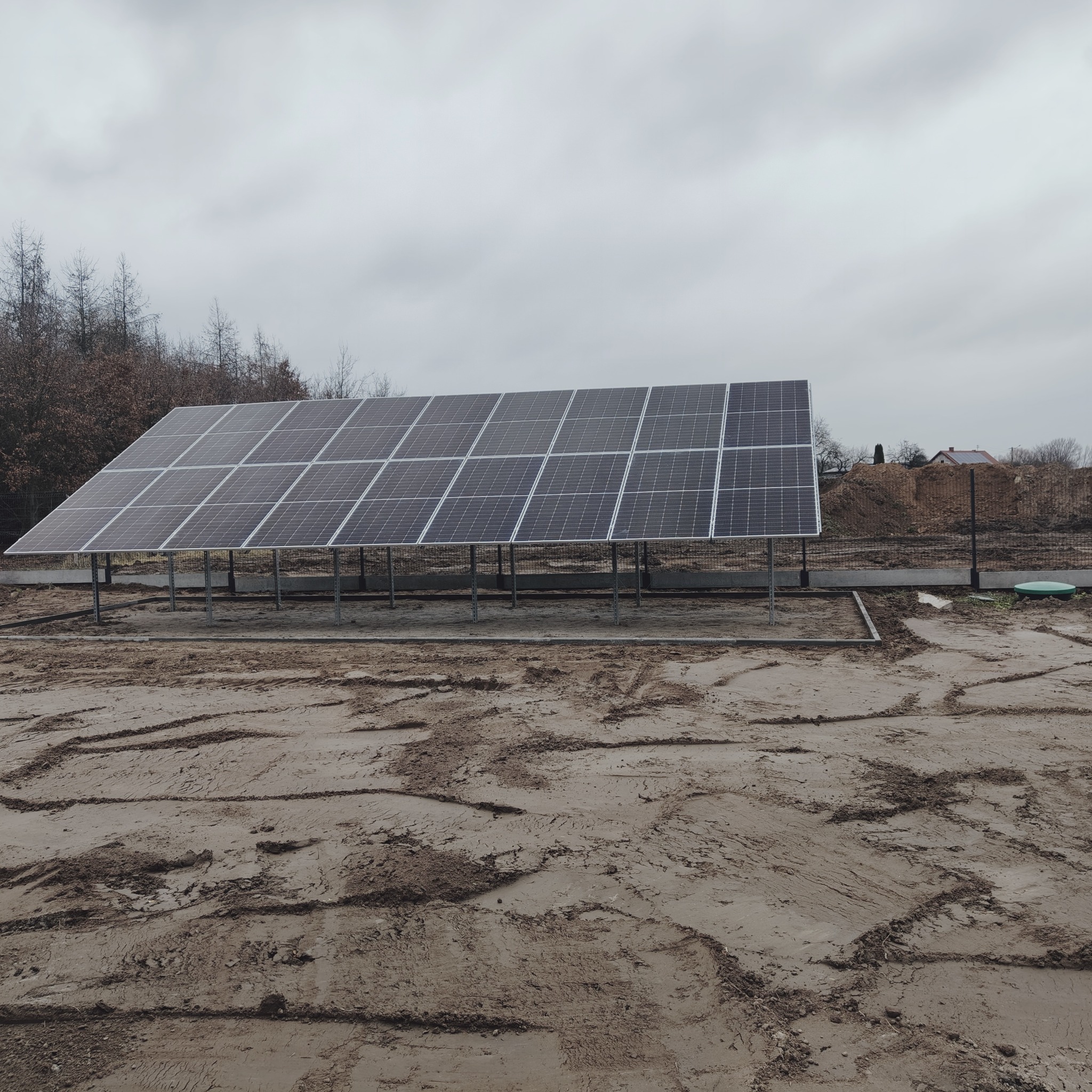 Rząd paneli słonecznych na metalowej konstrukcji, na ubitej ziemi z widocznymi śladami opon, w tle drzewa i fragment budynku w pochmurny dzień.