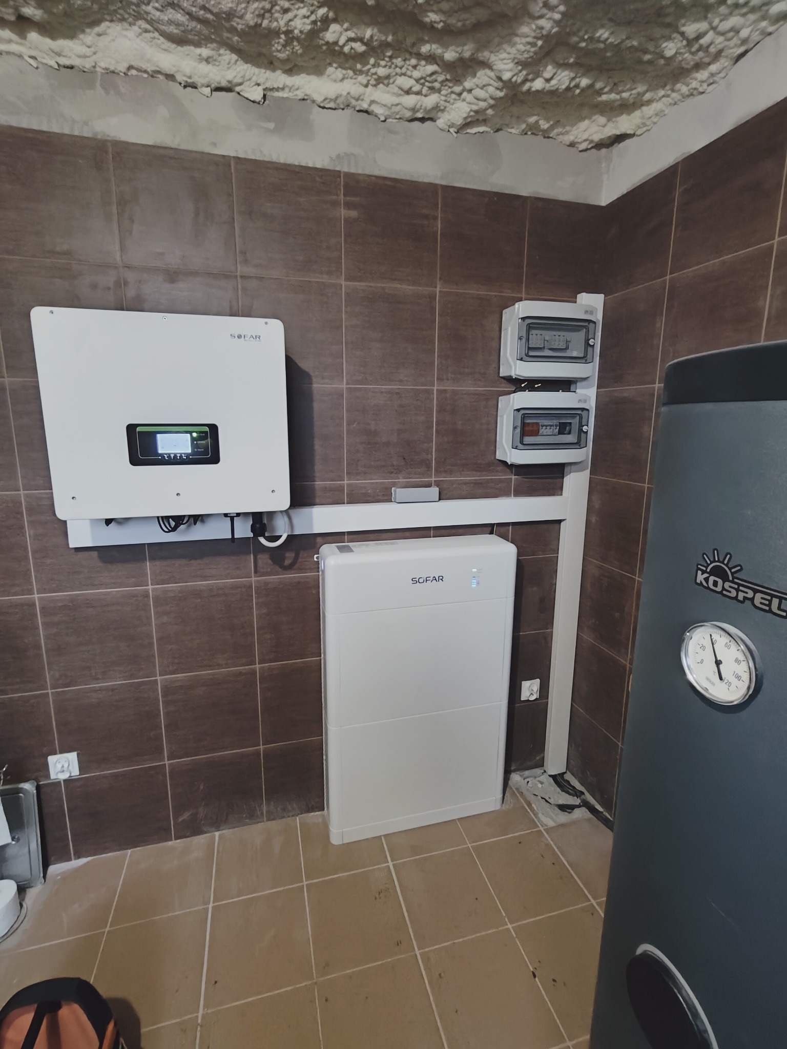 Biała bateria magazynu energii Sofar i falownik fotowoltaiczny tej samej marki zamontowane na brązowych płytkach ściennych w pomieszczeniu z wykończonym, ale niepomalowanym sufitem, obok szary...