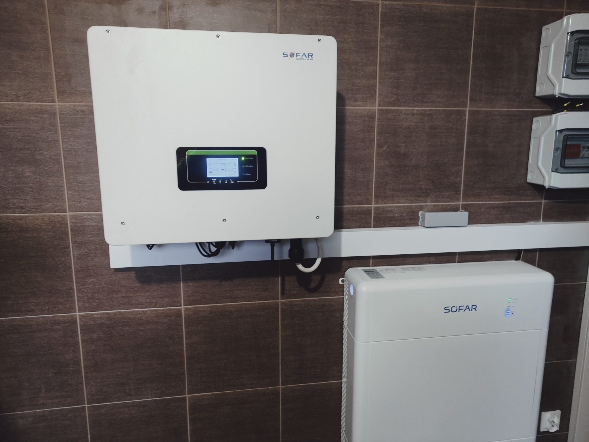 Biały inwerter i akumulator firmy SOFAR zamontowane na brązowej, kafelkowanej ścianie, obok liczników elektrycznych.