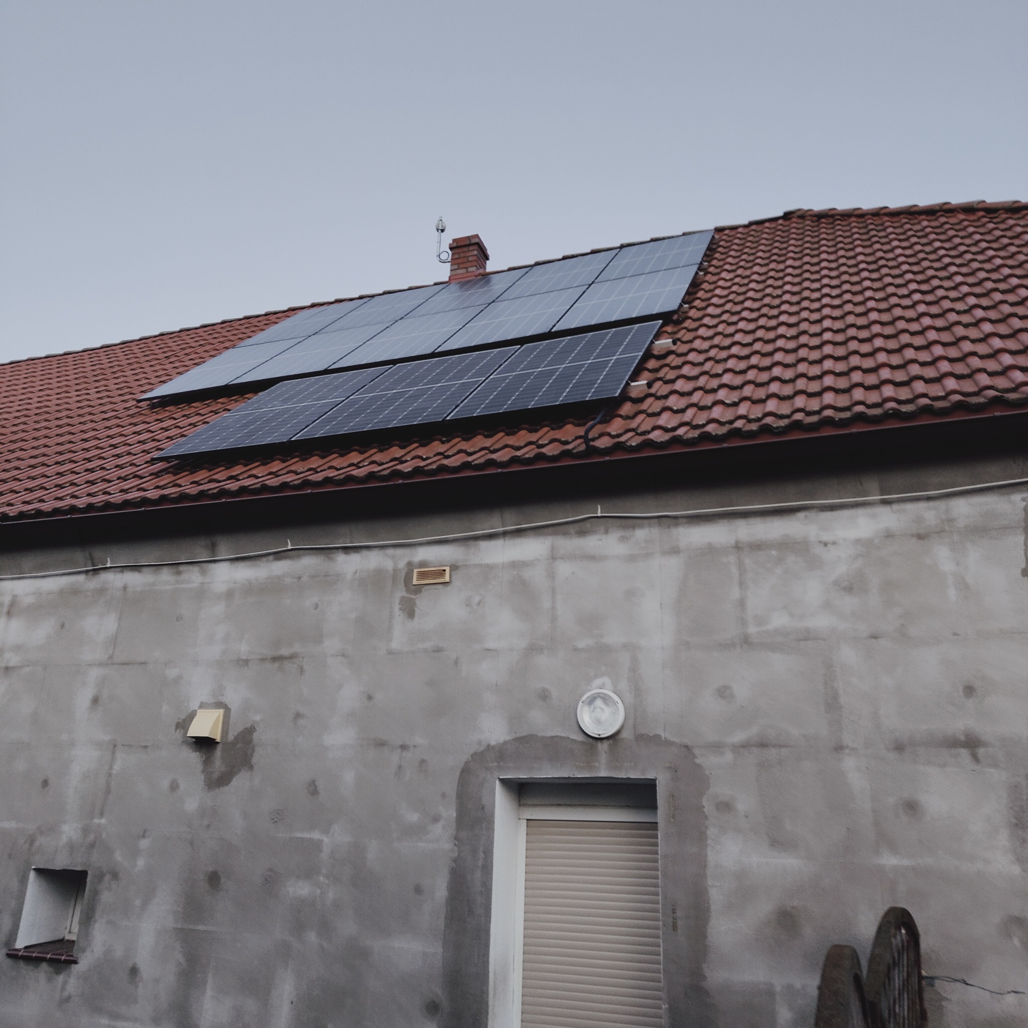 Dwa rzędy paneli słonecznych na czerwonej, ceramicznej dachówce starszego budynku z szarą, surową elewacją. Widoczny komin i antena.