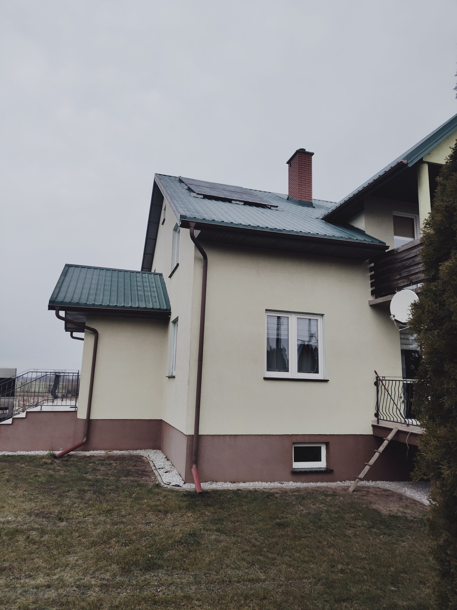 Dom jednorodzinny z panelami słonecznymi na dachu pokrytym blachodachówką w kolorze zielonym, widok z boku na tle pochmurnego nieba.