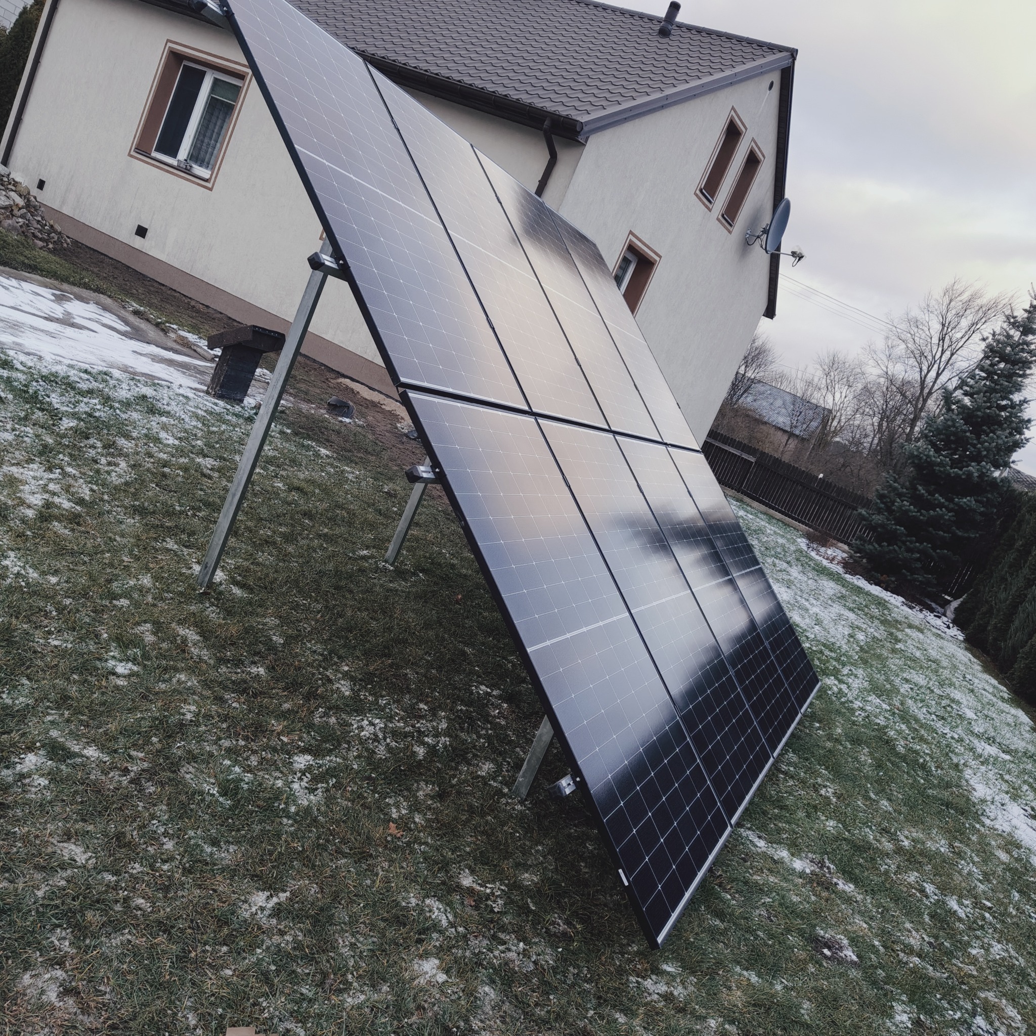 Cztery panele fotowoltaiczne na metalowej konstrukcji, ustawione na trawniku z resztkami śniegu, na tle domu jednorodzinnego z anteną satelitarną.