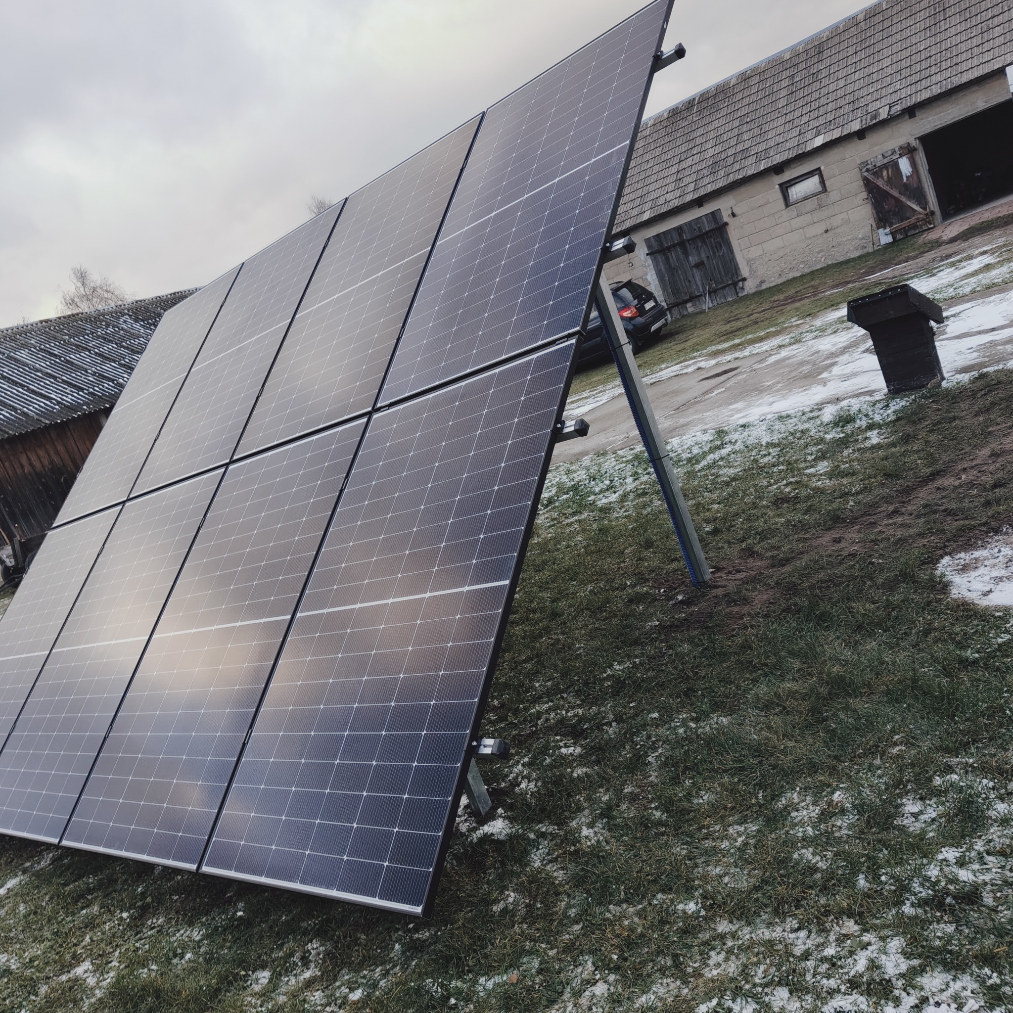 Panel fotowoltaiczny składający się z kilku modułów, ustawiony na trawniku przed budynkiem gospodarczym z widocznym samochodem w tle, częściowo pokrytym śniegiem.