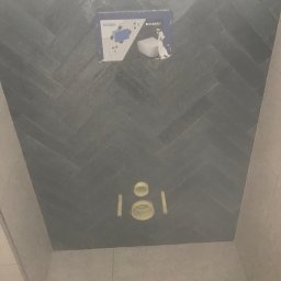Bau best - Ściana łazienki wykończona szarą glazurą ułożoną we wzór jodełki, z widoczną instalacją podtynkową Geberit Sigma do WC, przed zabudową stelaża i montażem ceramiki.