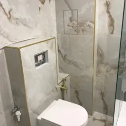 Wykończona łazienka z wiszącą toaletą, marmurowymi płytkami z brązowymi żyłami i złotymi listwami dekoracyjnymi.