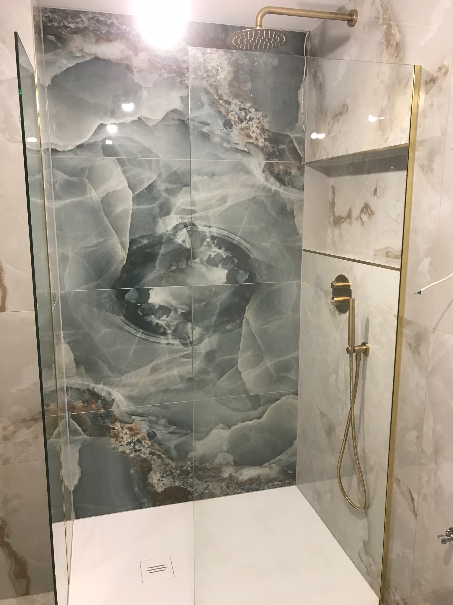 Kabina prysznicowa z marmurowymi płytkami w odcieniach szarości i beżu, złota armatura, deszczownica i odpływ liniowy w białym brodziku.