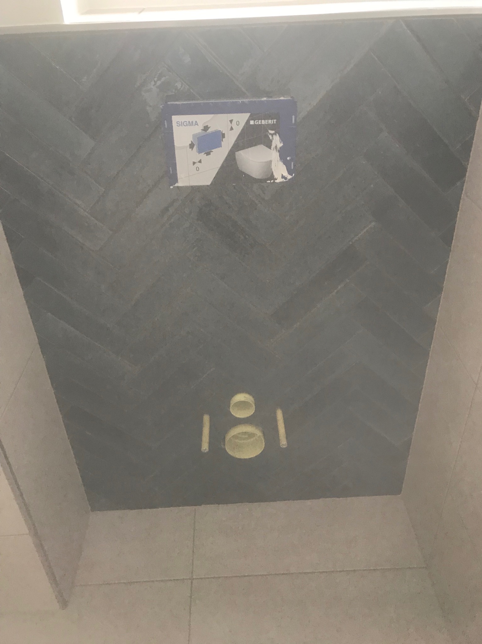 Ściana łazienki wykończona szarą glazurą ułożoną we wzór jodełki, z widoczną instalacją podtynkową Geberit Sigma do WC, przed zabudową stelaża i montażem ceramiki.