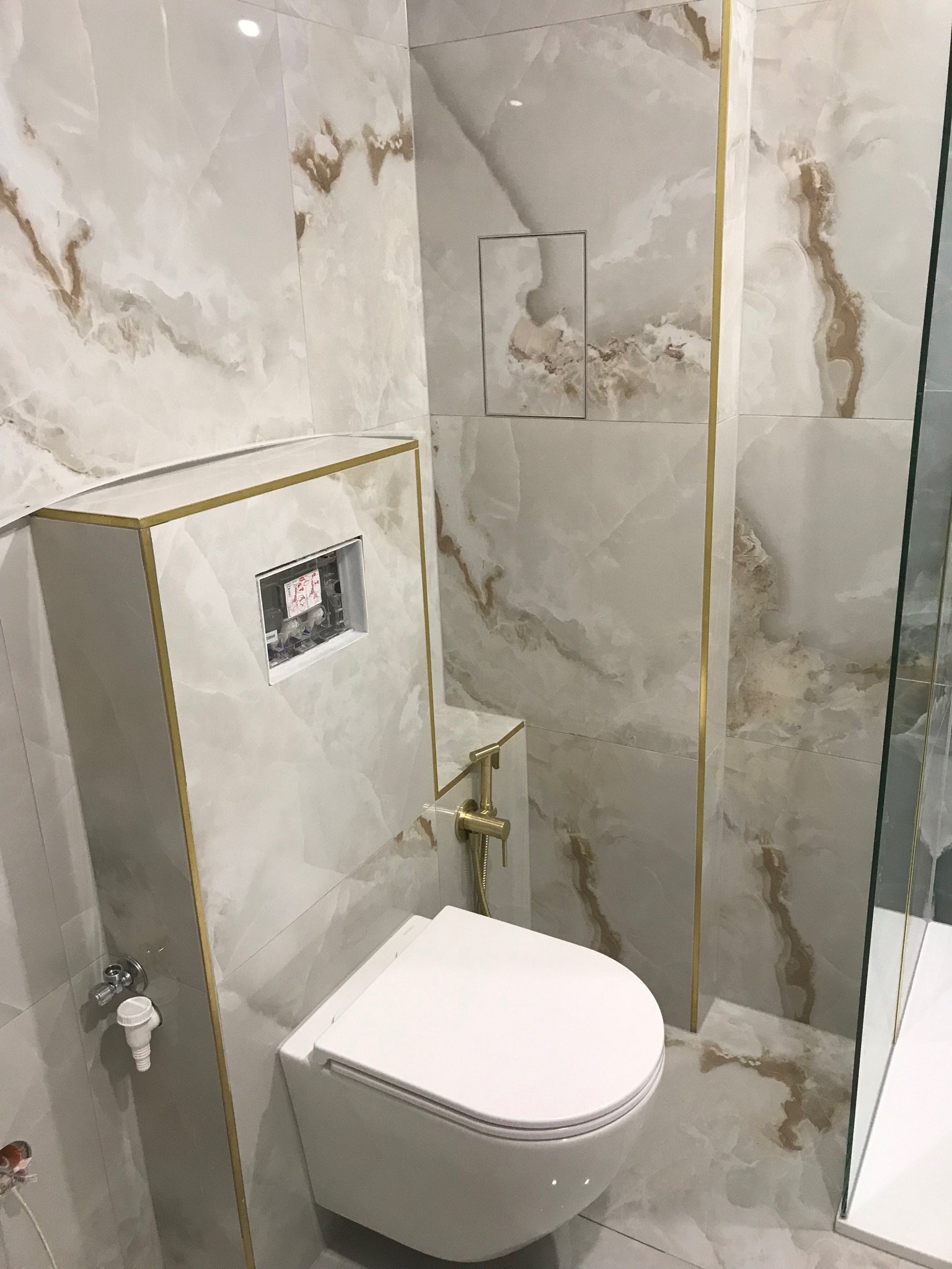 Wykończona łazienka z wiszącą toaletą, marmurowymi płytkami z brązowymi żyłami i złotymi listwami dekoracyjnymi.