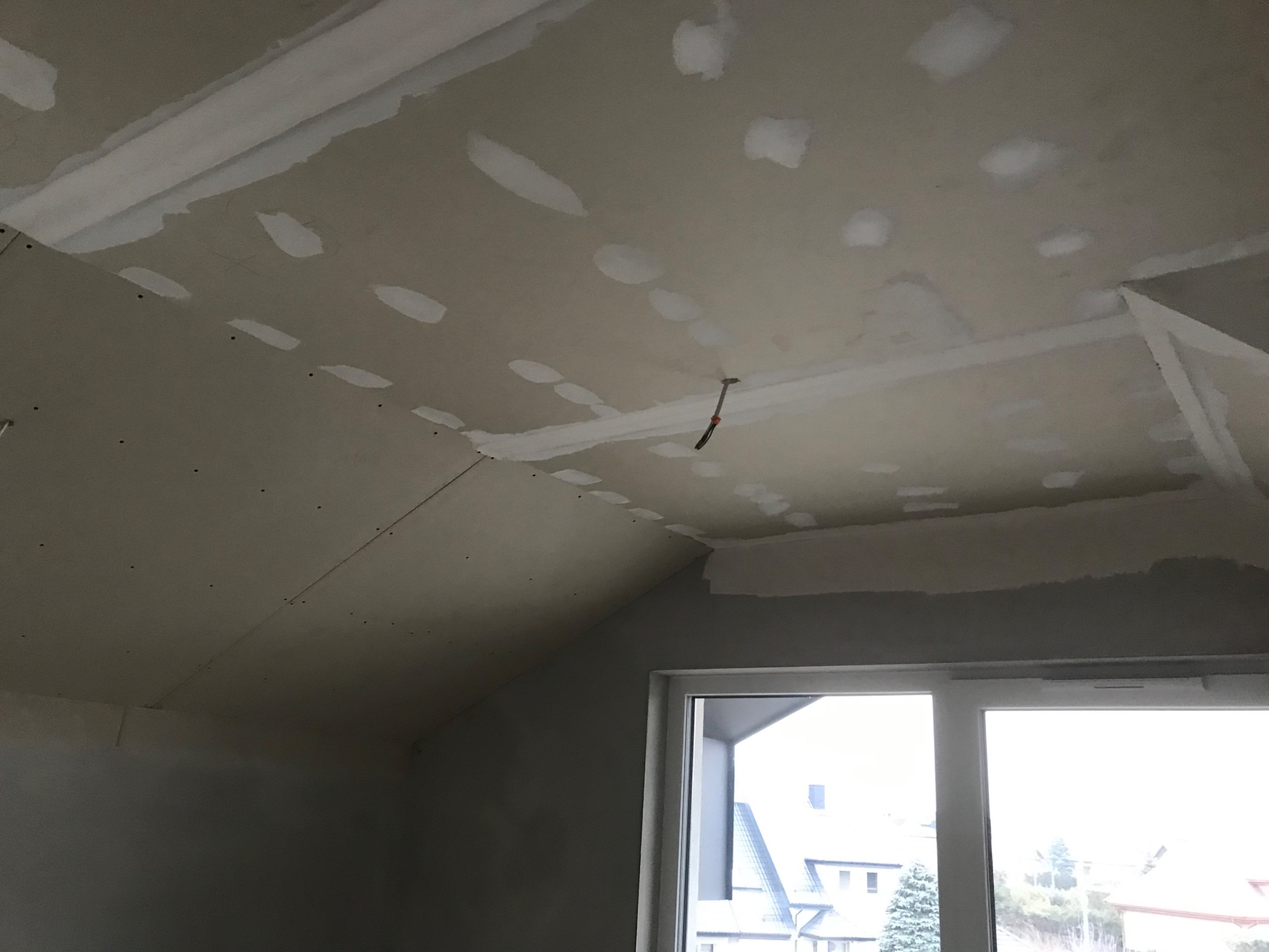 Poddasze w trakcie wykańczania: widok na sufit z zamontowaną płytą gipsowo-kartonową, szpachlowane łączenia i wkręty, wystający kabel elektryczny, okno z widokiem na zabudowę mieszkalną.